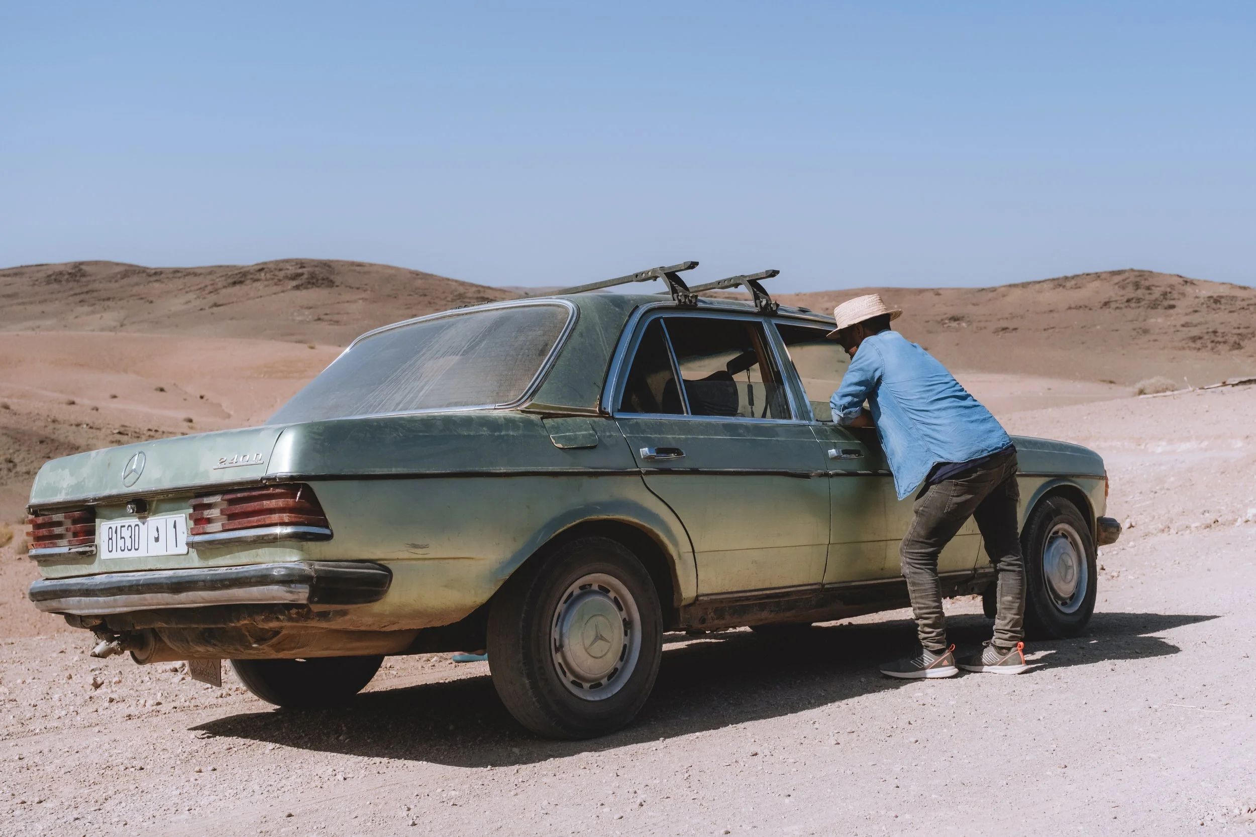 Une personne regarde à l'intérieur d'une voiture Mercedes-Benz vintage en désert avec des dunes en arrière-plan.