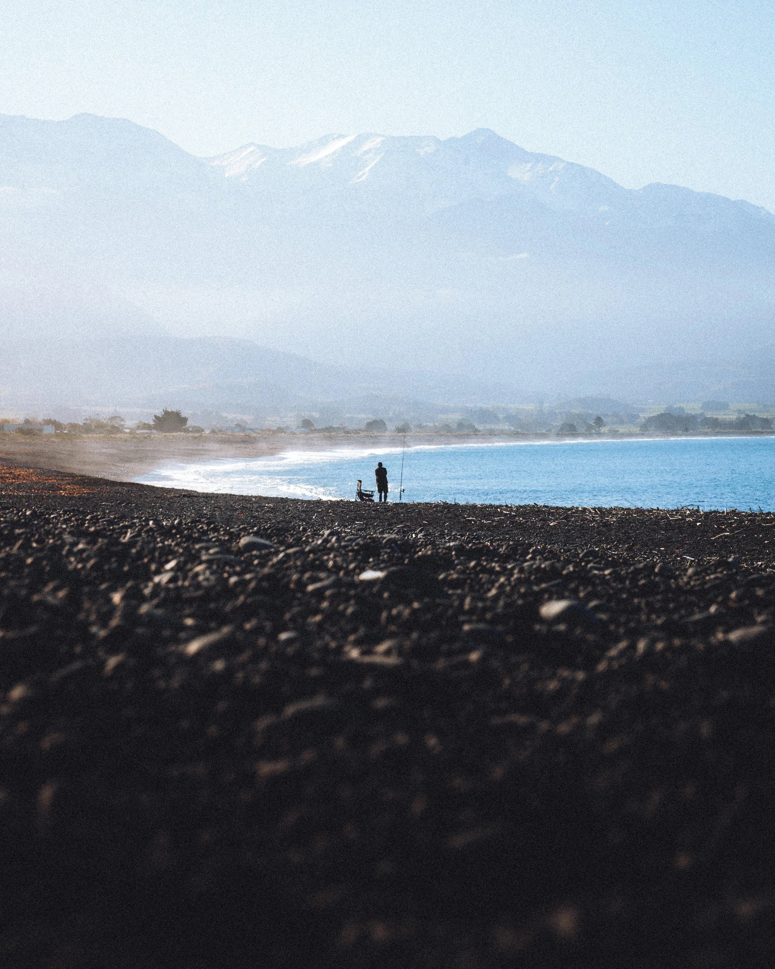Une personne pêchant sur une plage rocheuse avec une montagne enneigée en arrière-plan.