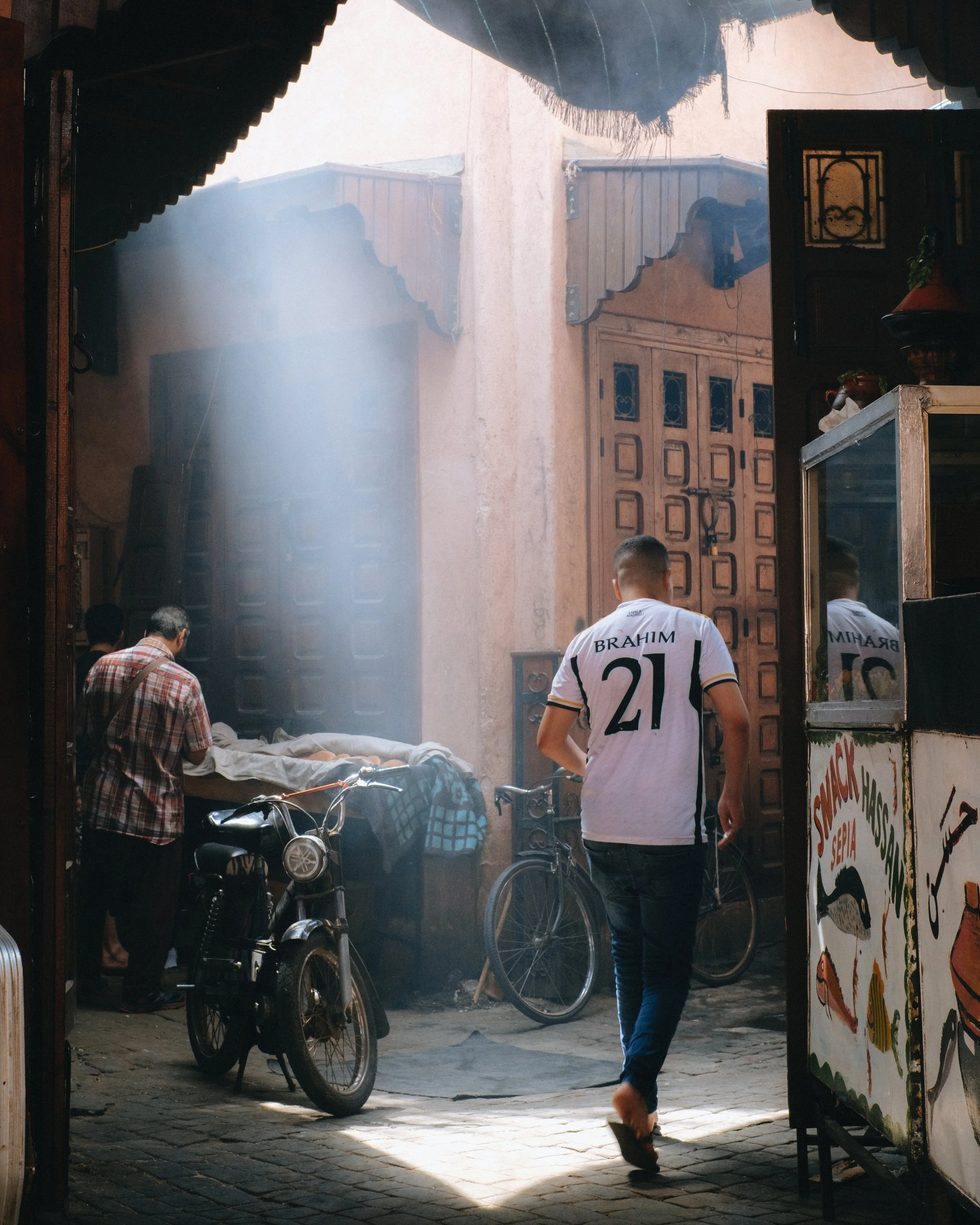 Une scène en rue avec deux personnes, un homme portant un maillot de football blanc avec le nom 'Brahim' et le numéro 21, et un autre homme en chemise à carreaux. Il y a aussi des vélos, un stand de nourriture, une porte en bois, et un peu de lumière