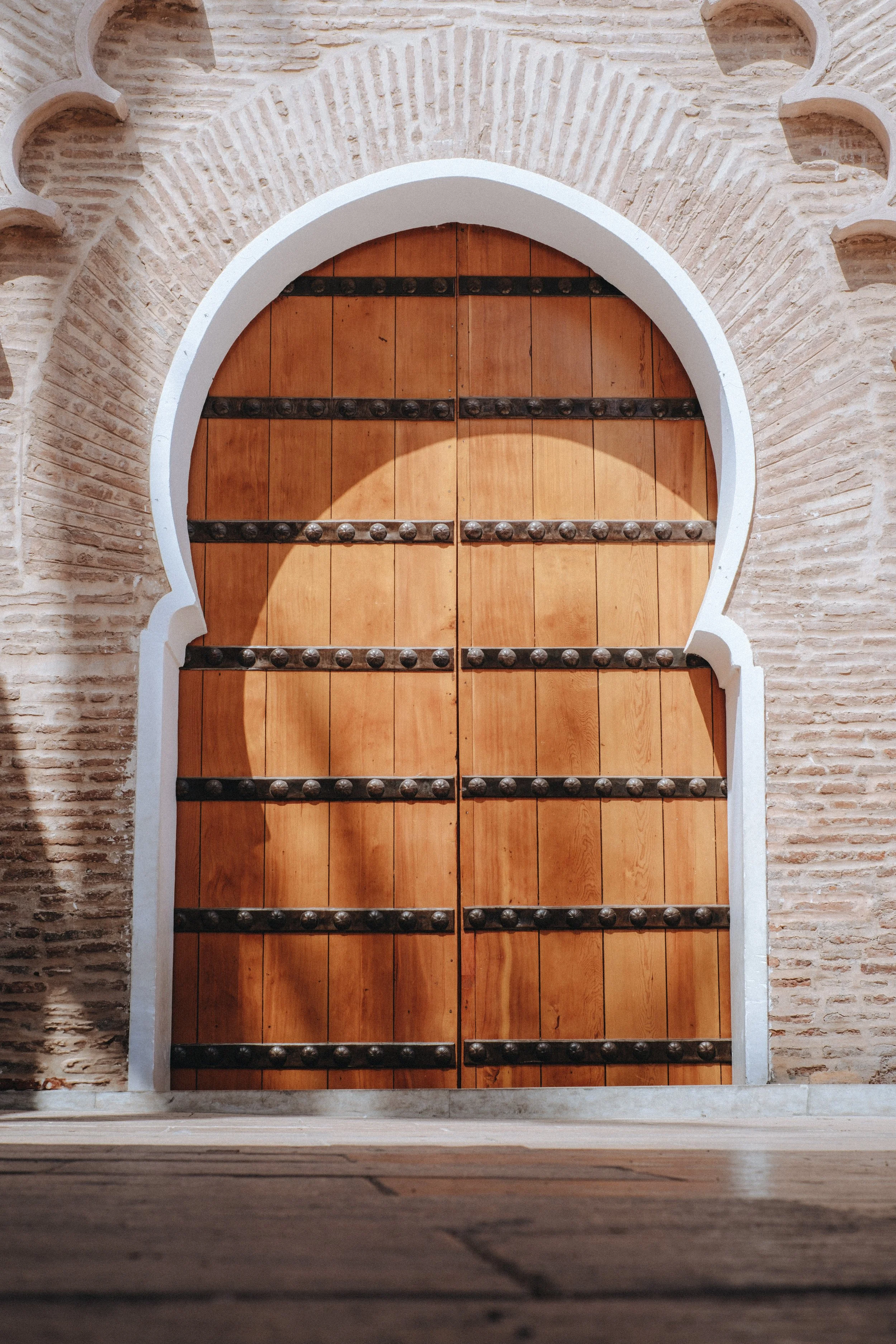 Une grande porte en bois avec des barres métalliques, encadrée par une arche blanche en pierre, faisant partie d'un mur en briques beige, typique de l’architecture ancienne ou méditerranéenne.