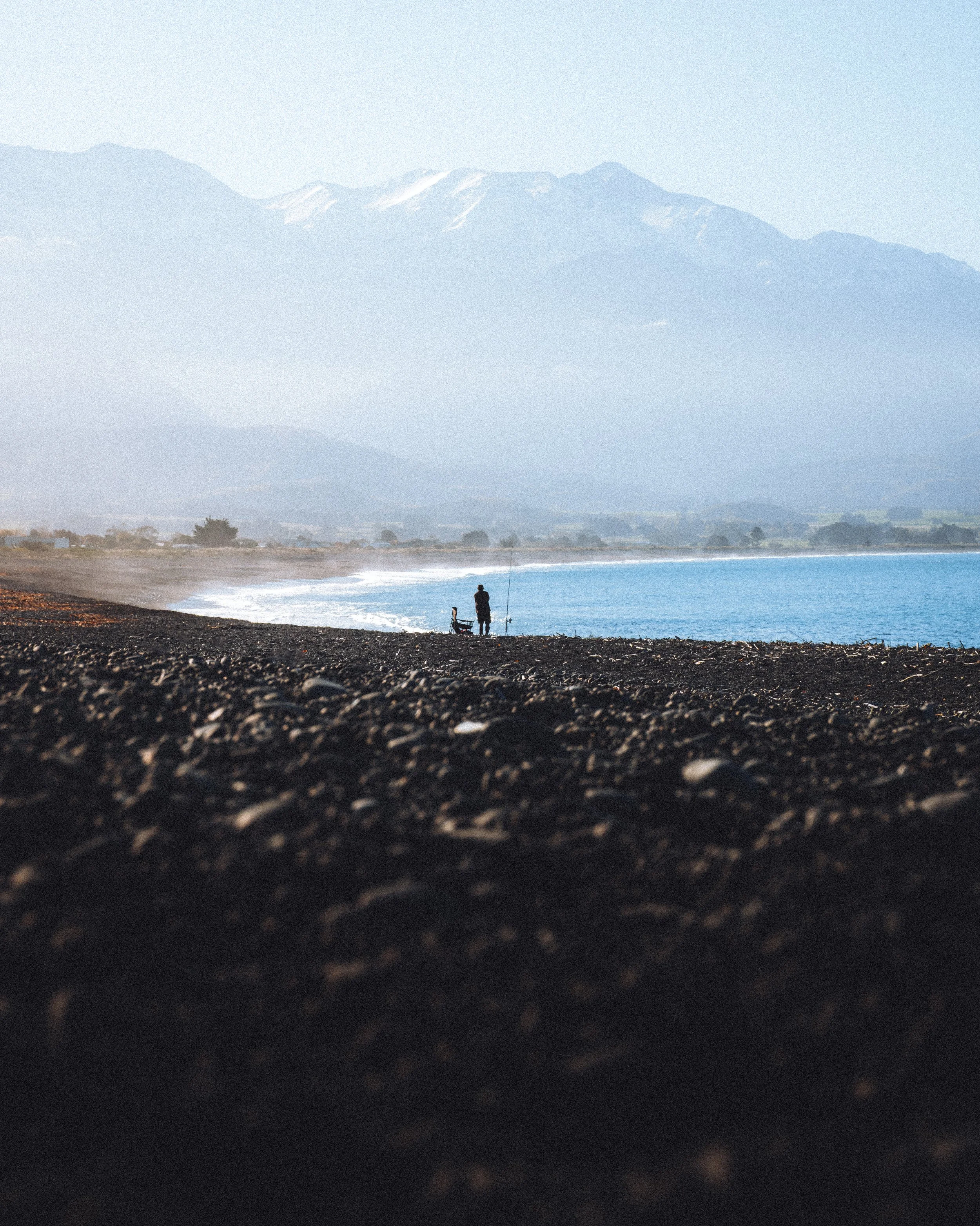 Un pêcheur seul sur une plage de galets, avec des montagnes en arrière-plan et la mer à ses côtés.