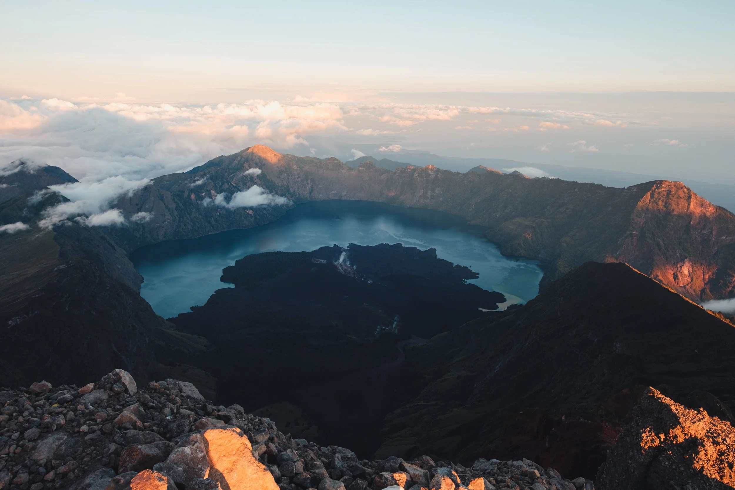 Vue aérienne d'un volcan avec un lac calde dans le cratère, entouré par des montagnes et des nuages, lors du lever ou coucher du soleil.