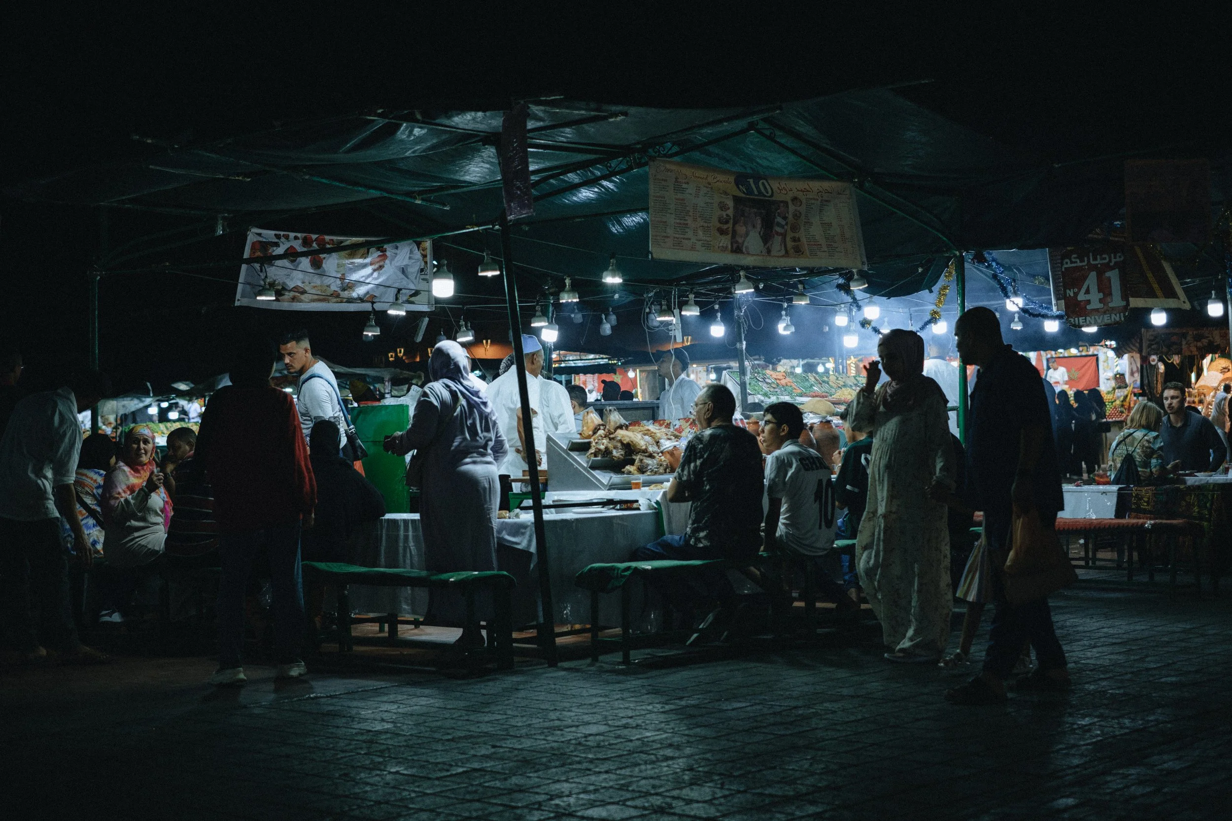 Marché nocturne avec vendeurs et clients, étals éclairés, cuisine et produits alimentaires visibles, ambiance animée et animée.