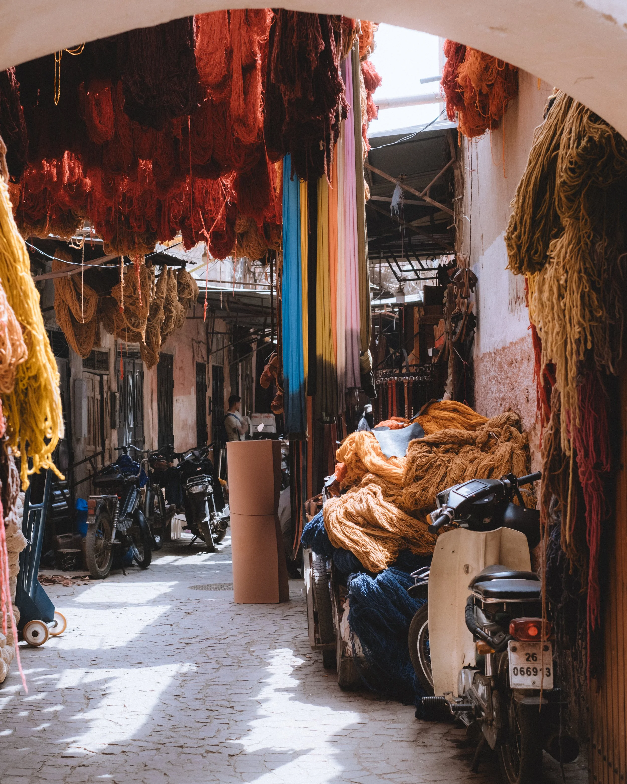 Rue étroite avec des tissus colorés et des motos garées, illuminée par la lumière naturelle du soleil.
