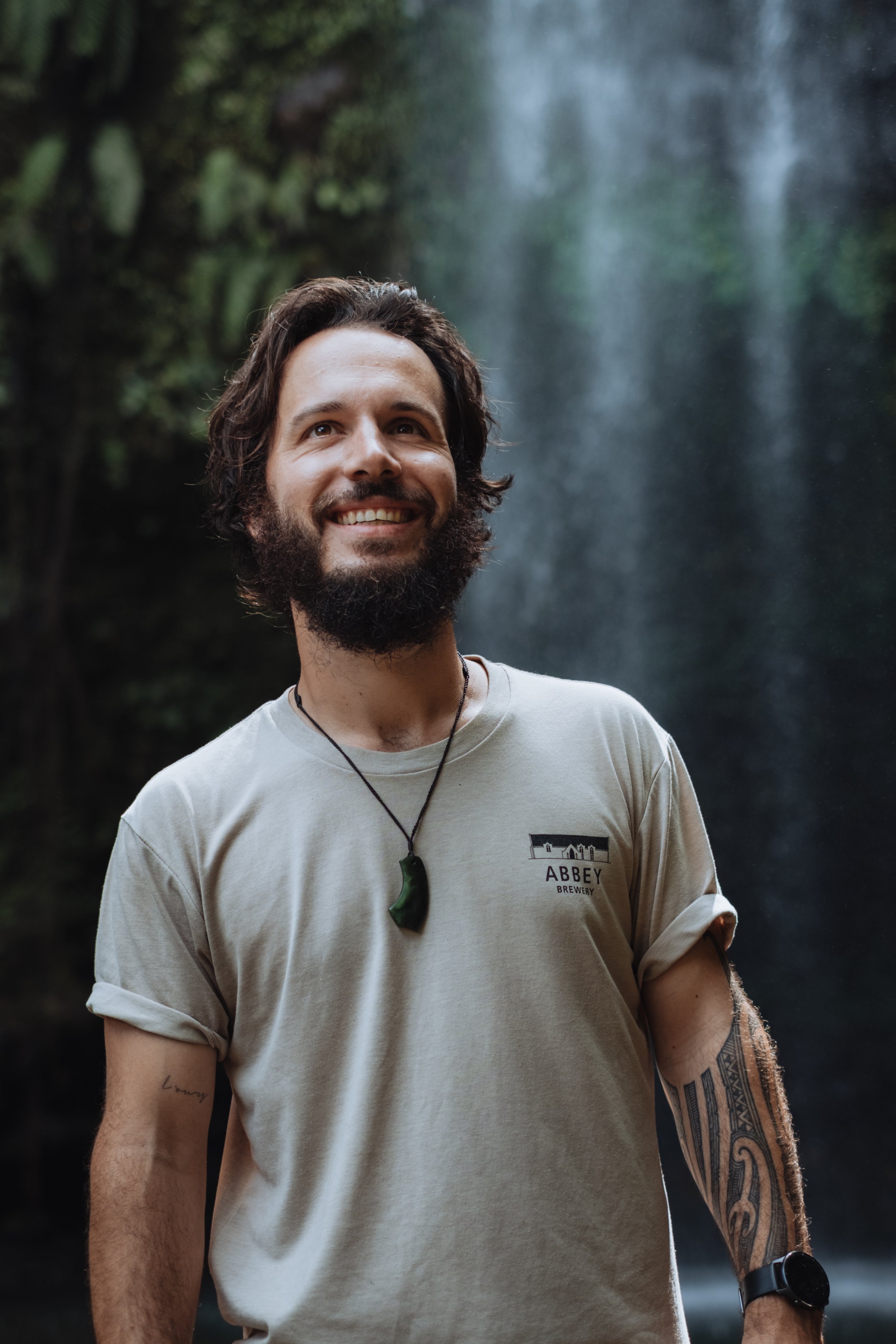 Homme souriant avec barbe, porte un t-shirt beige avec logo d'une brasserie, un collier avec pendentif vert, en nature devant une cascade.