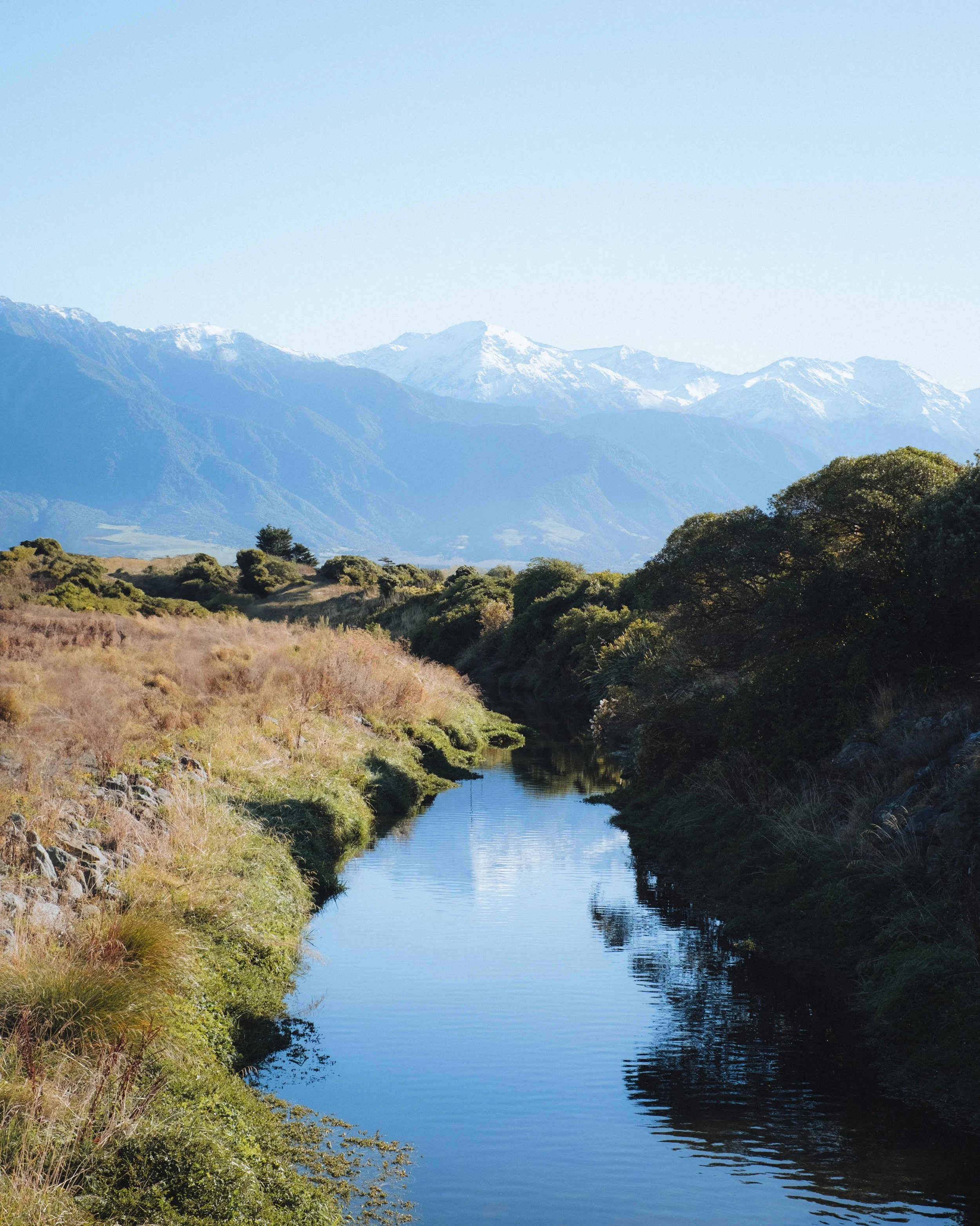 Vue d'une rivière serpentant à travers un paysage avec des montagnes enneigées en arrière-plan, des arbustes et de l'herbe verte sur les rives.