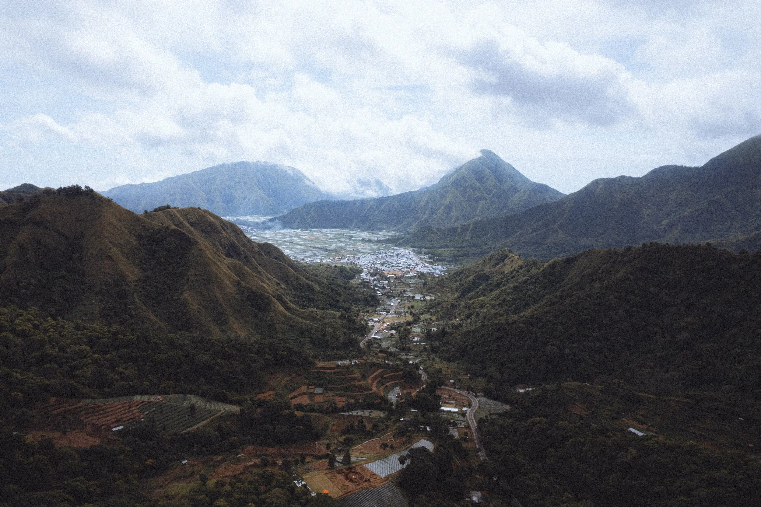 Paysage de montagnes verdoyantes avec une vallée, des champs agricoles et un village au centre, sous un ciel partiellement nuageux.