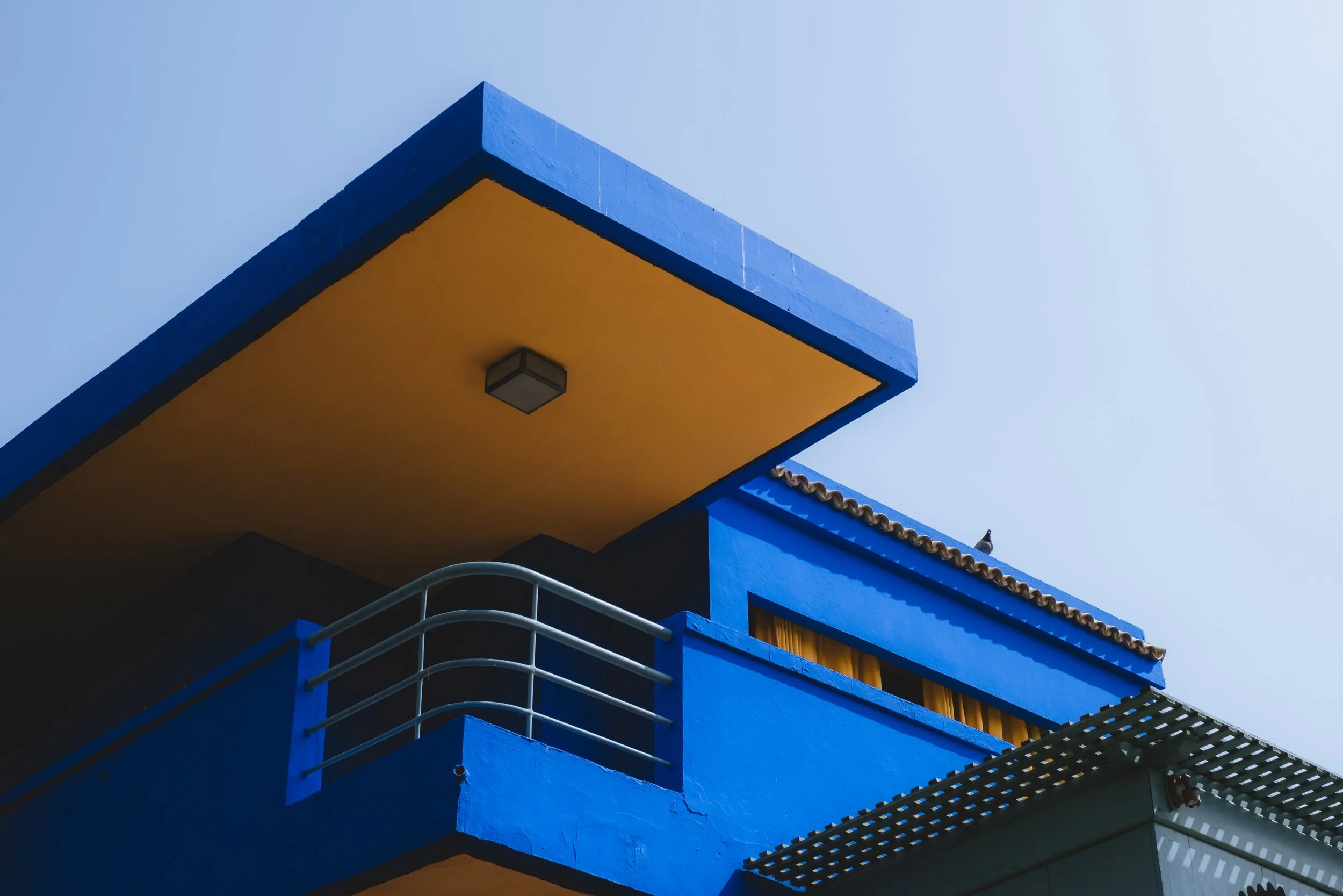 Vue d'un bâtiment moderne peint en bleu avec une surélévation jaune, un balcon avec une barrière métallique, un toit en tuiles et un ciel clair en arrière-plan.
