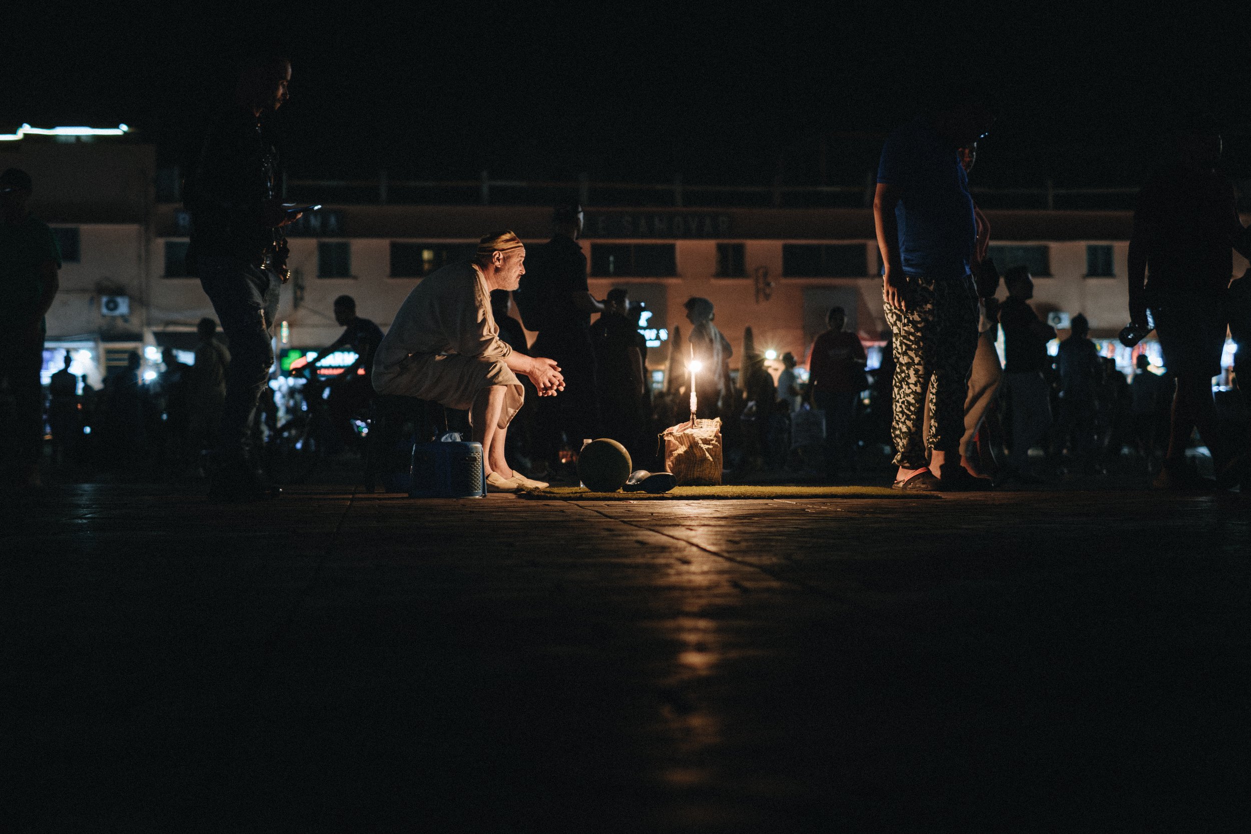 Une scène nocturne dans un lieu public avec plusieurs personnes. Au centre, une personne assise près d'une bougie allumée, entourée d'objets. D'autres individus se tiennent ou se déplacent, certains dans l'ombre, dans une atmosphère sombre avec des l