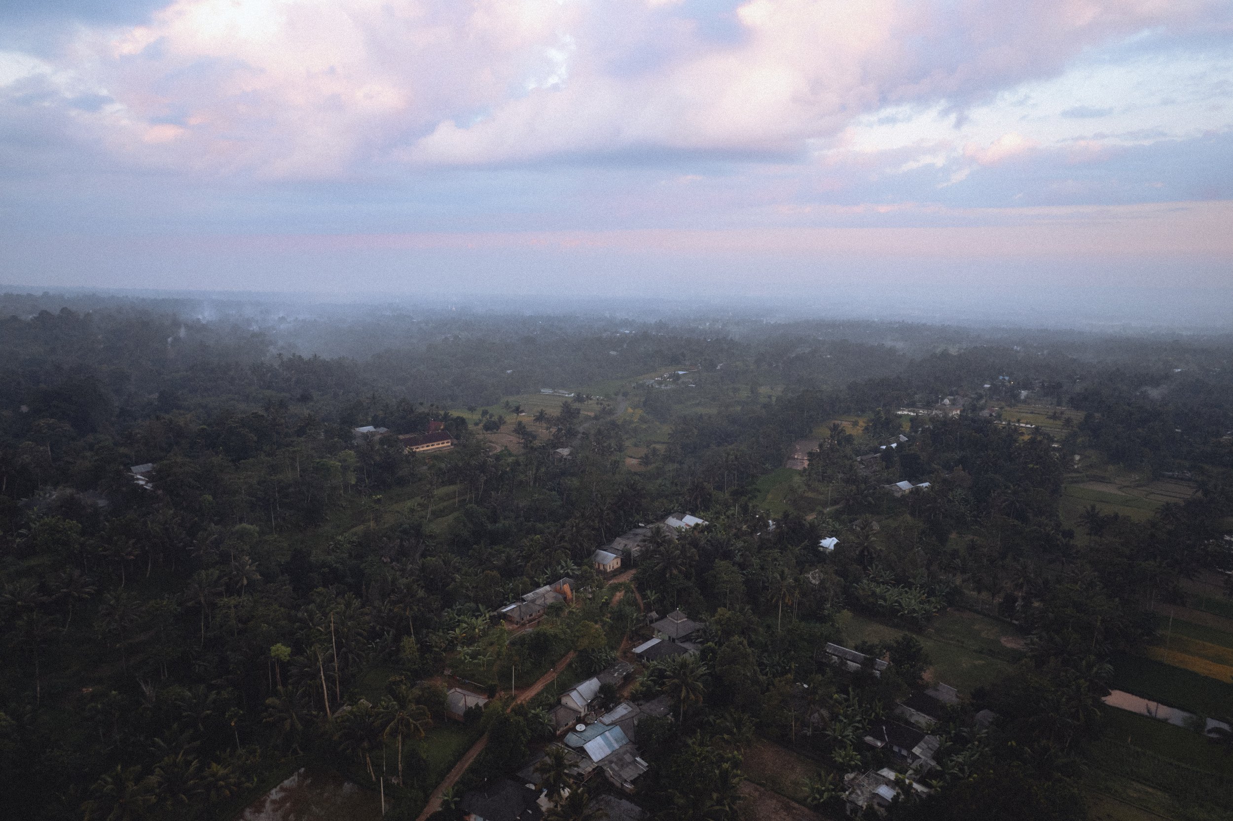 Vue aérienne d'une zone rurale avec des champs, des maisons et des arbres sous un ciel nuageux au coucher du soleil.