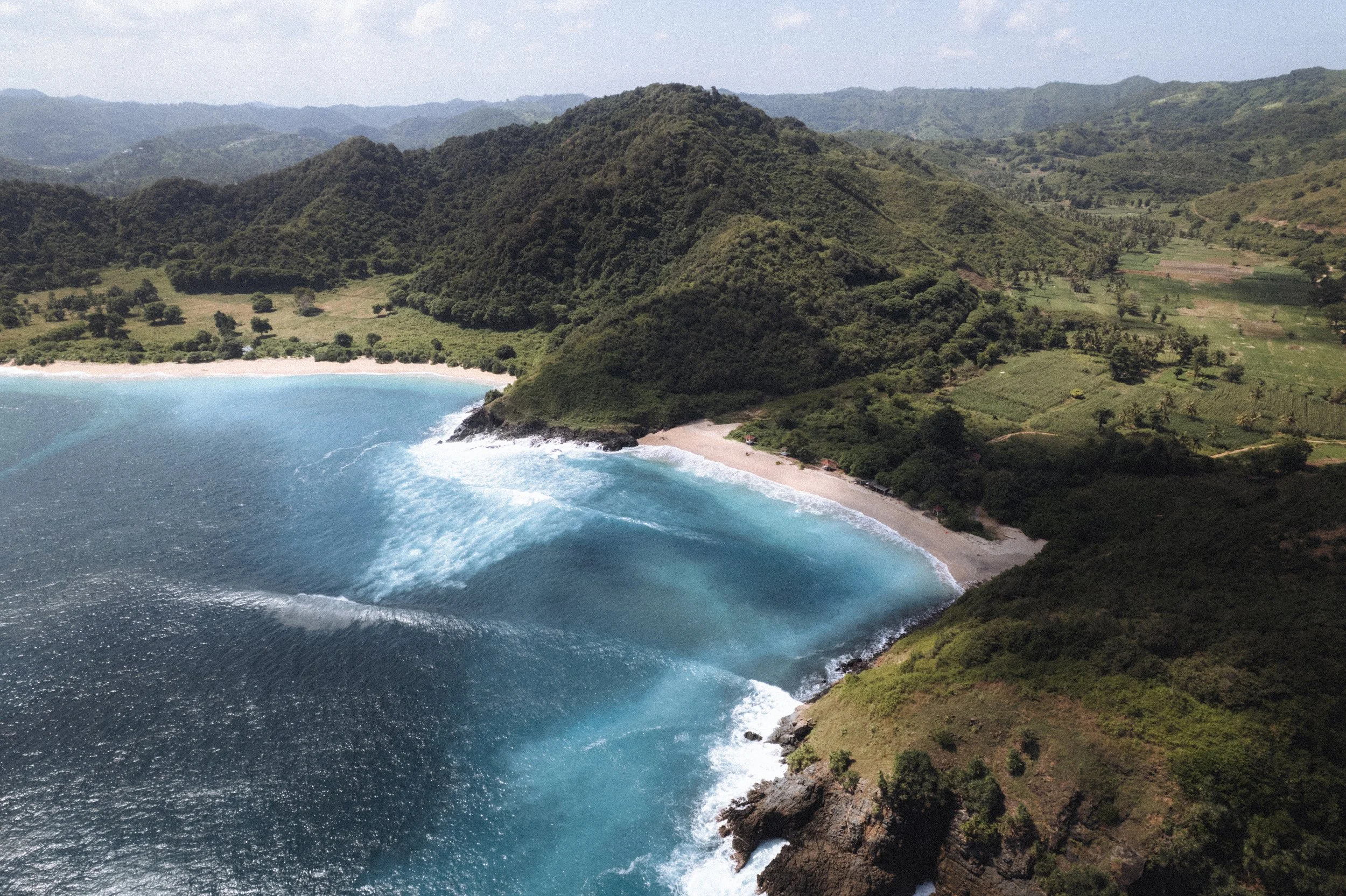 Une plage de sable blanc bordée par une mer bleue et entourée de collines verdoyantes et montagneuses.