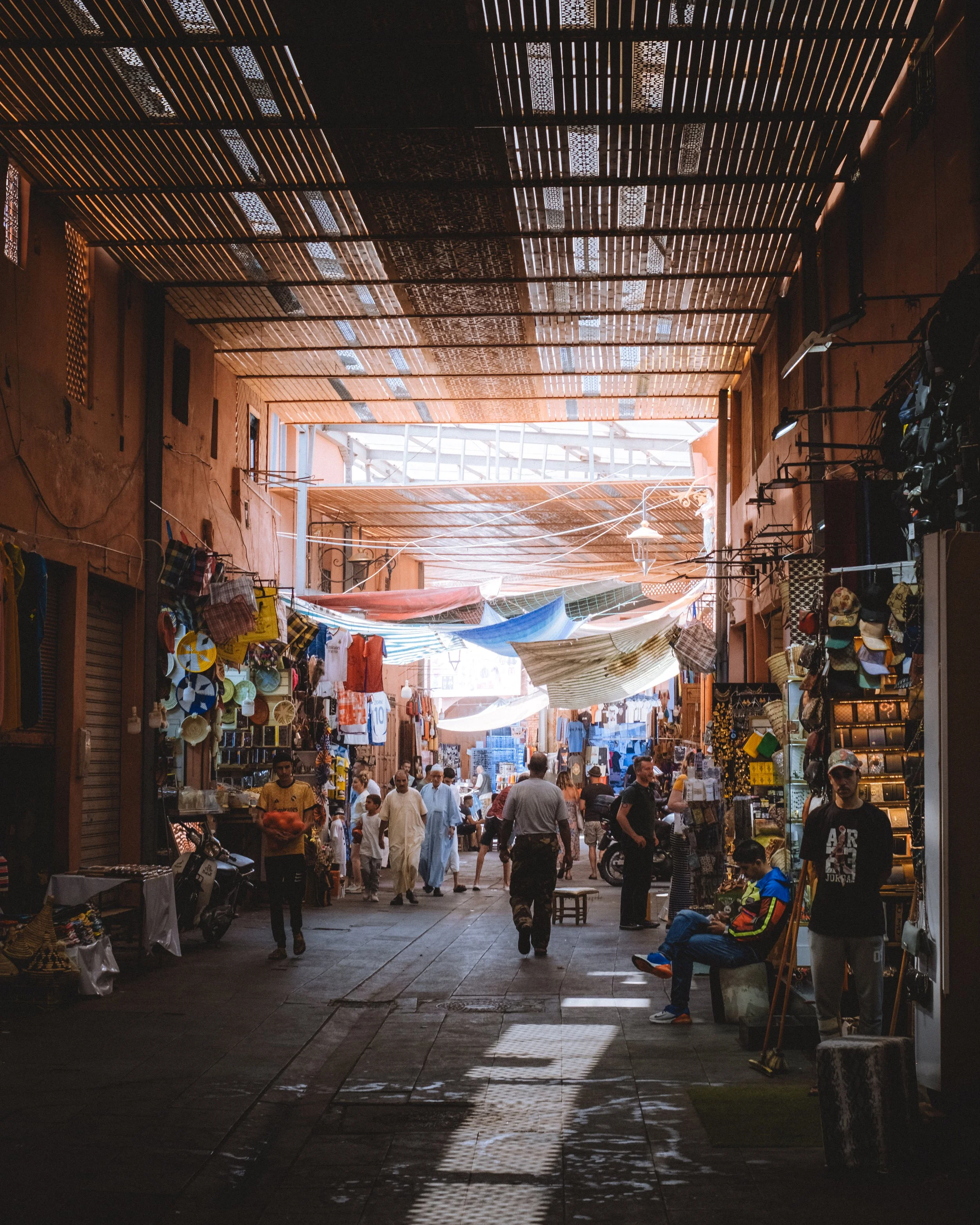 Marché couvert avec des vendeurs et des acheteurs, étals avec des marchandises, tissu suspendu au plafond, ambiance animée et ombragée.