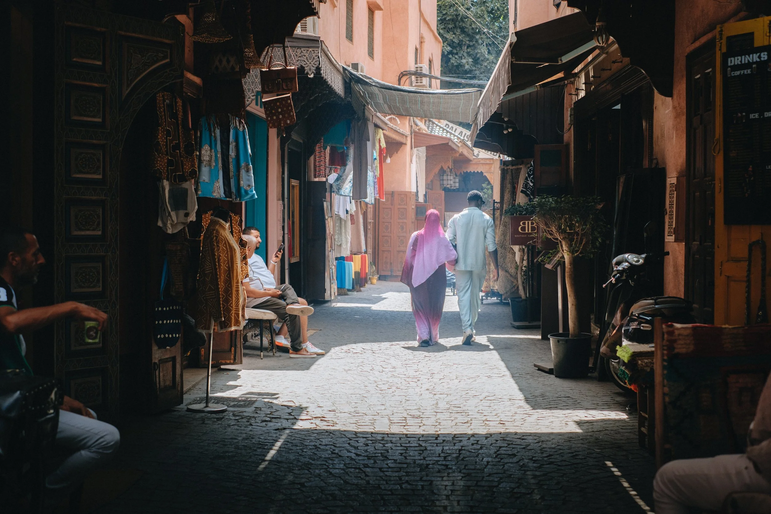 Une rue commerçante en médina, avec des boutiques de vêtements et de souvenirs, des personnes assises sous les abris, et un couple marchant par la rue en train de discuter.