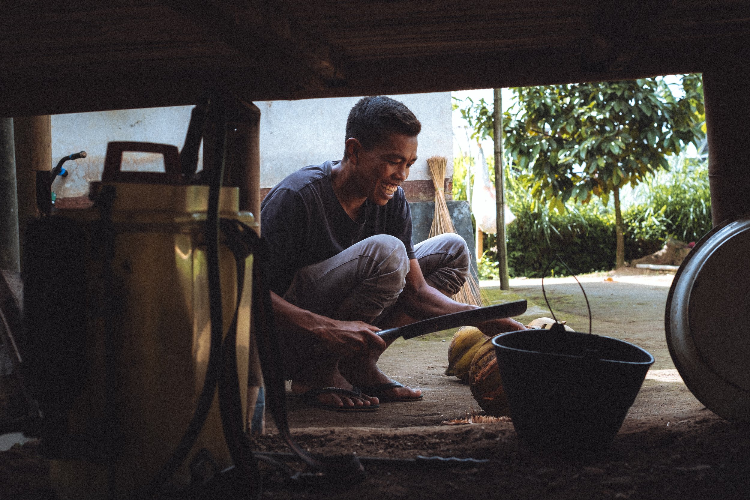 Jeune homme souriant, coupant une noix de coco avec une machette dans une maisonnette en plein air, entouré de végétation.