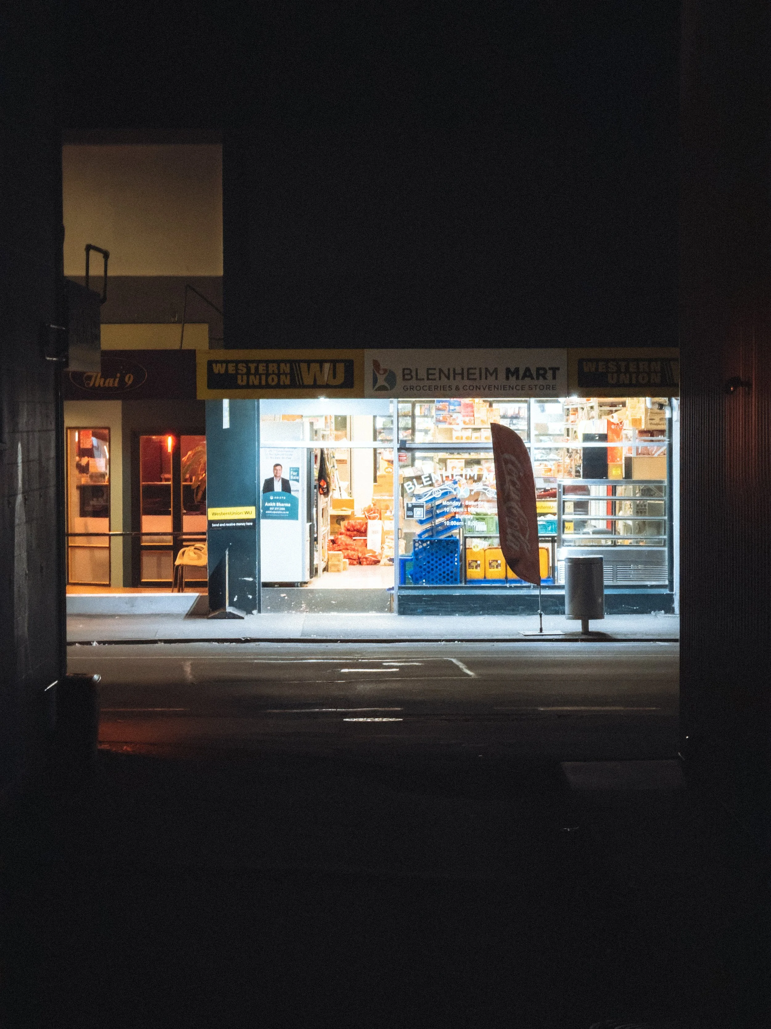 L'entrée d'un supermarché éclairée la nuit, avec une enseigne indiquant 'Blenheim Mart' et des logos de Western Union, contenant des marchandises visibles à l'intérieur.