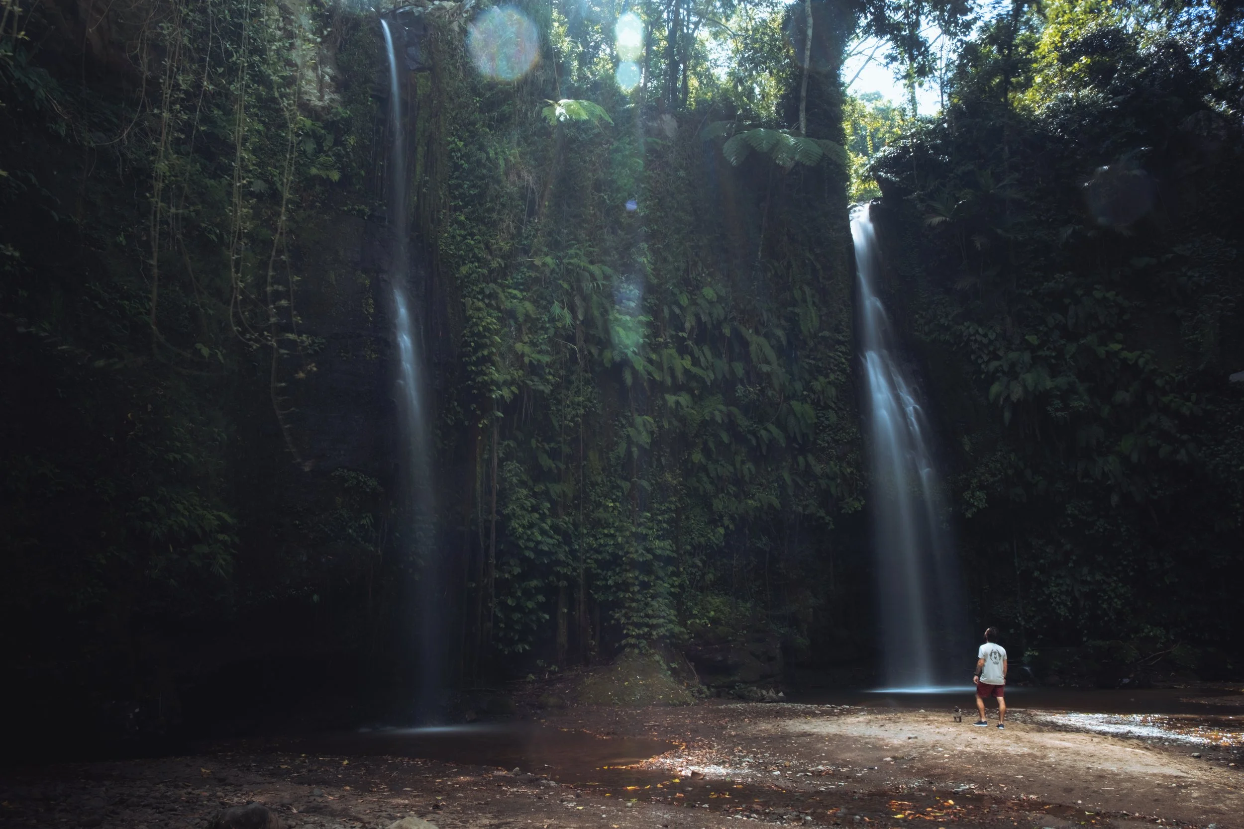 Une personne debout devant deux cascades dans une forêt tropicale dense, avec de la végétation luxuriante et de la lumière du soleil traversant les arbres.