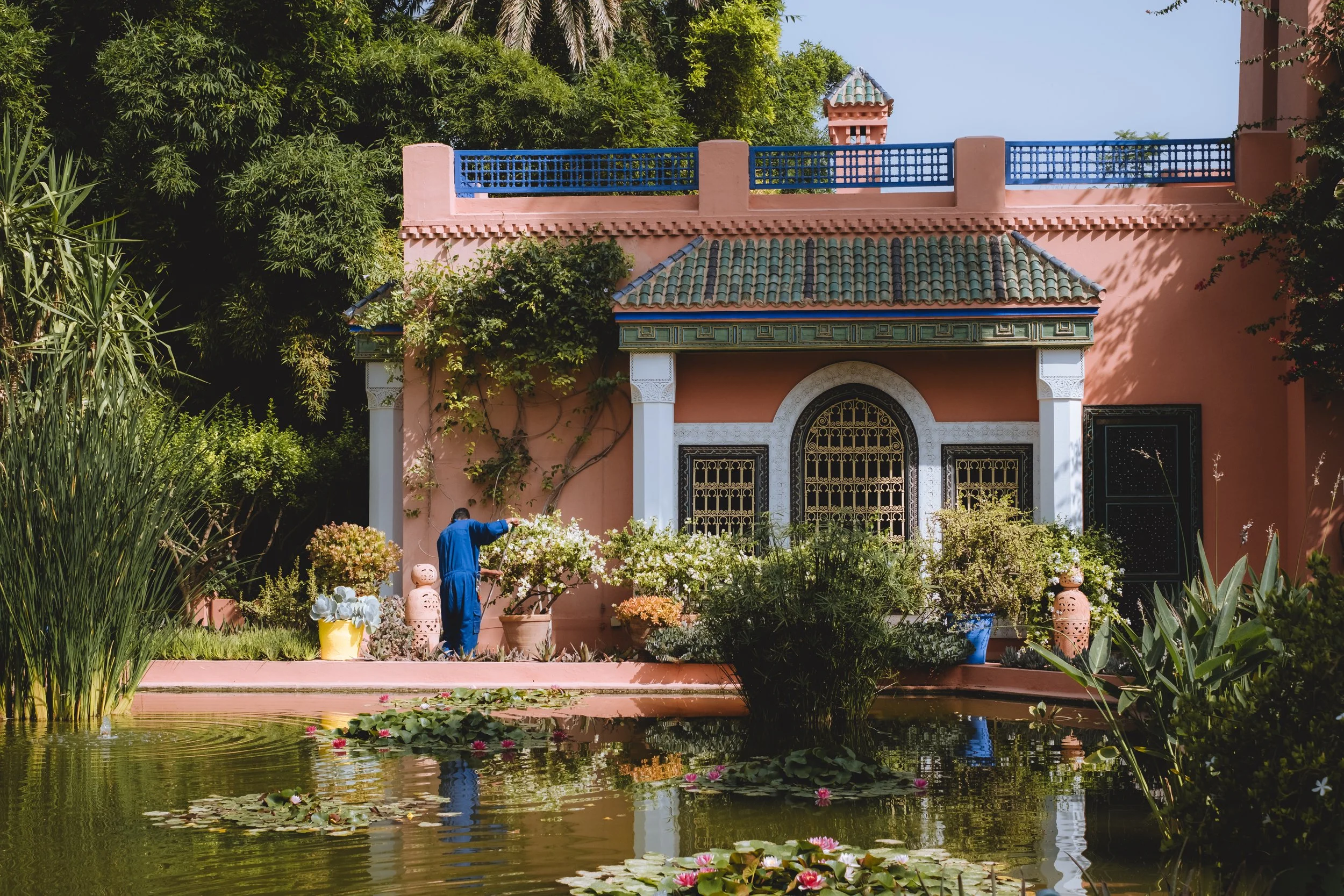 Une maison de style méditerranéen rose avec un toit en tuiles, entourée de verdure et d'un jardin avec un étang contenant des nénuphars, et un homme en bleu arrosant des plantes.