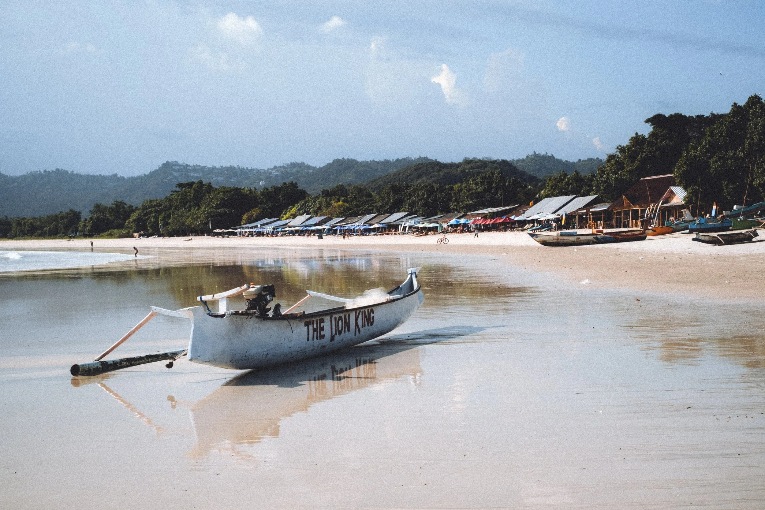 Bateau blanc nommé 'The Lion King' sur la plage, avec des parasols et des cabanes en bois en arrière-plan, forêt et montagnes au loin.