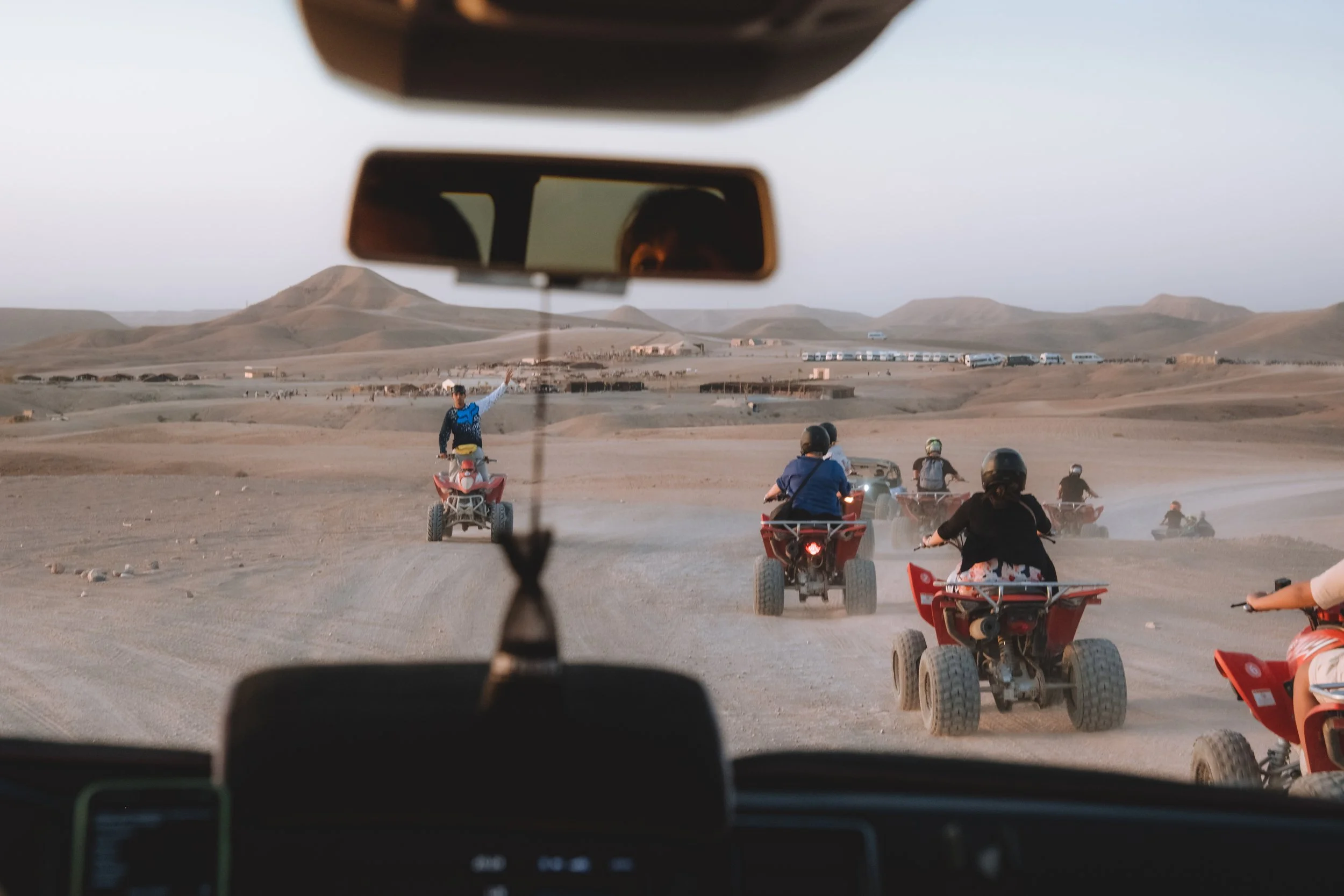 Groupe de personnes faisant du quad dans un désert avec des dunes et des montagnes à l'horizon, vue depuis l'intérieur d'un véhicule.