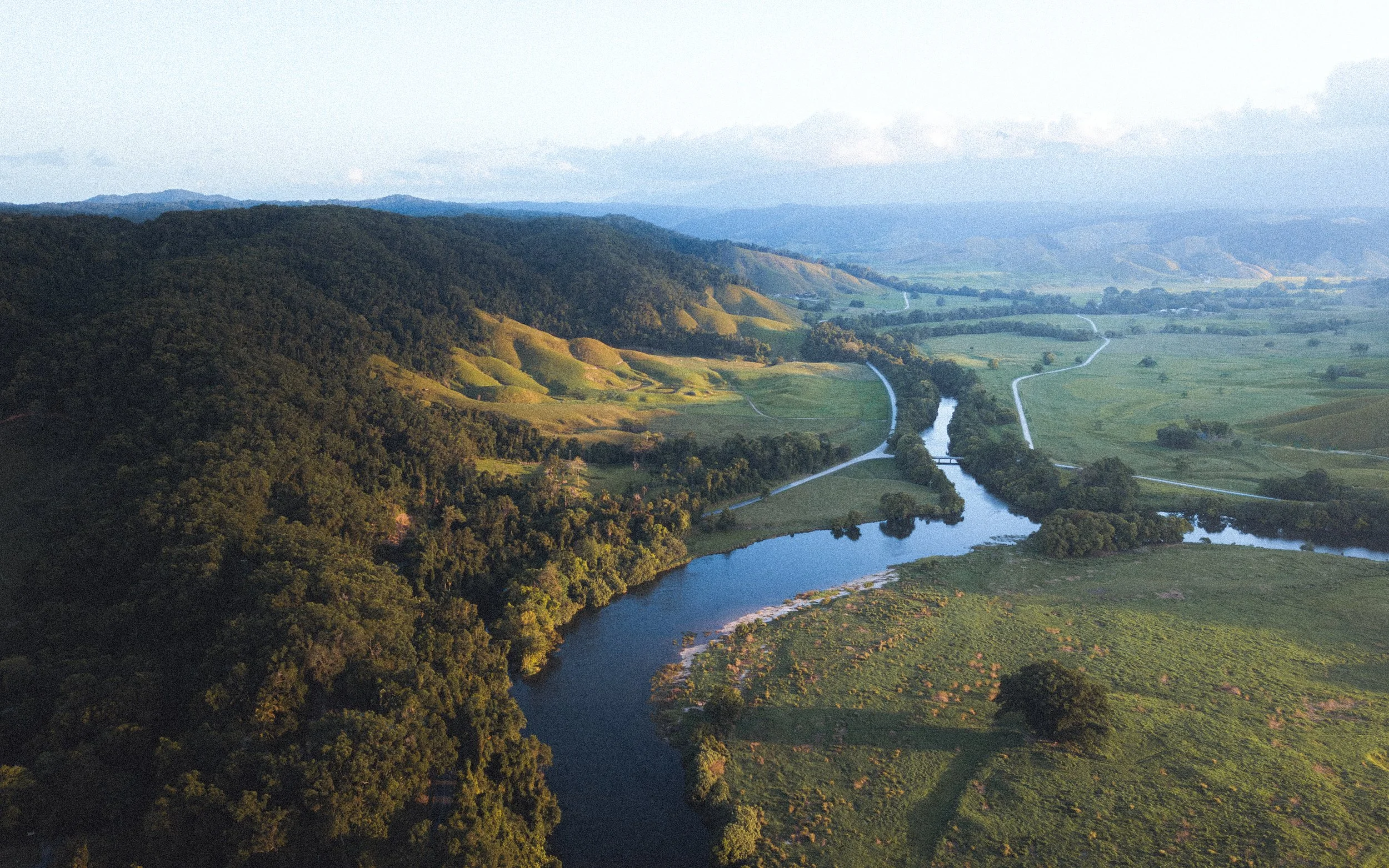 Vallée avec rivière sinueuse, champs verts, forêts et collines au loin lors d'une journée ensoleillée.