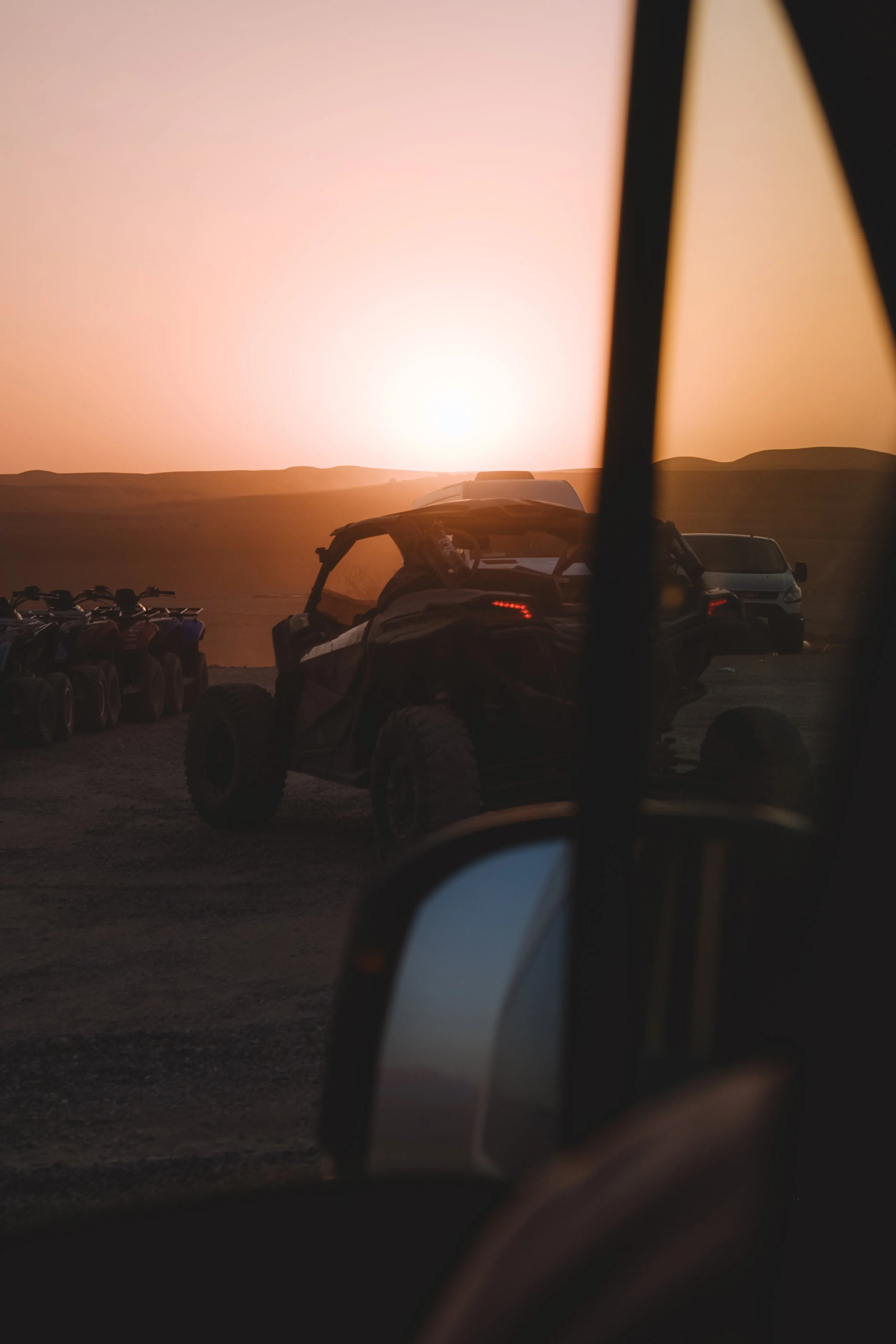 Coucher de soleil derrière un véhicule tout-terrain dans un désert, avec plusieurs autres véhicules tout-terrain et voitures en arrière-plan.