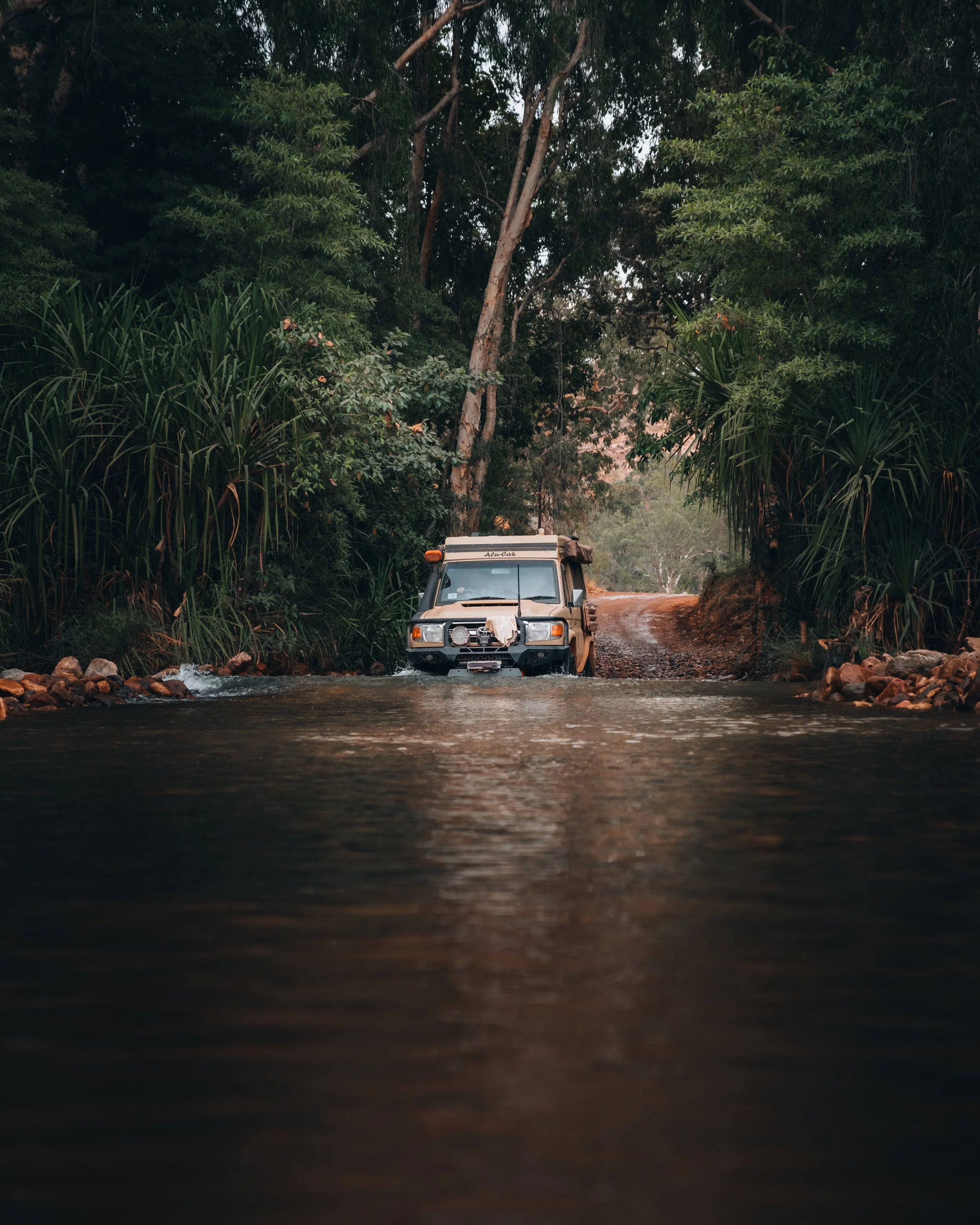 Un véhicule tout-terrain traverse une rivière dans une forêt dense avec des arbres et des arbustes verts. Gibb River Road, Australia