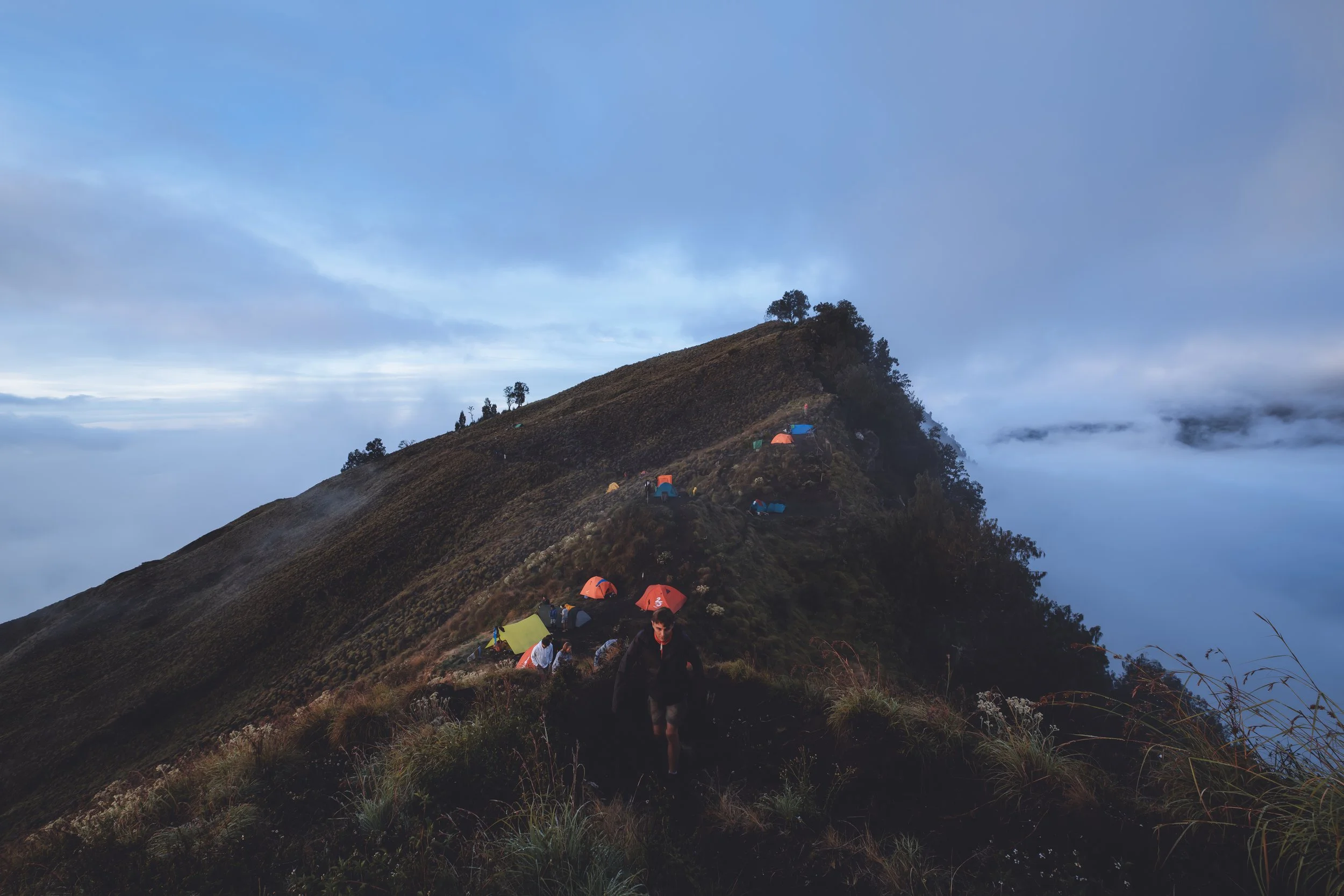 Un groupe de campeurs installés sur un sommet de montagne entouré de nuages, avec des tentes colorées, lors d'une excursion en montagne en fin d'après-midi ou au lever du soleil.
