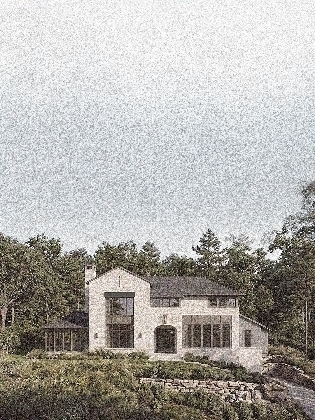 Large modern house with white exterior walls and multiple large windows, surrounded by trees and greenery, with a stone-lined pathway or landscaping in front.