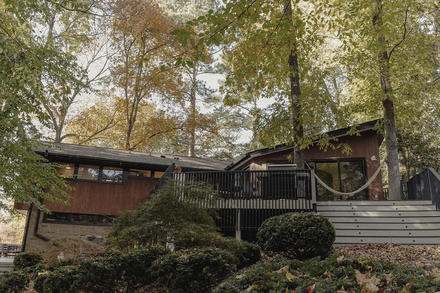 A modern house with large glass windows, a wooden exterior, and a deck with a hammock, surrounded by trees and shrubs in a wooded area during fall.