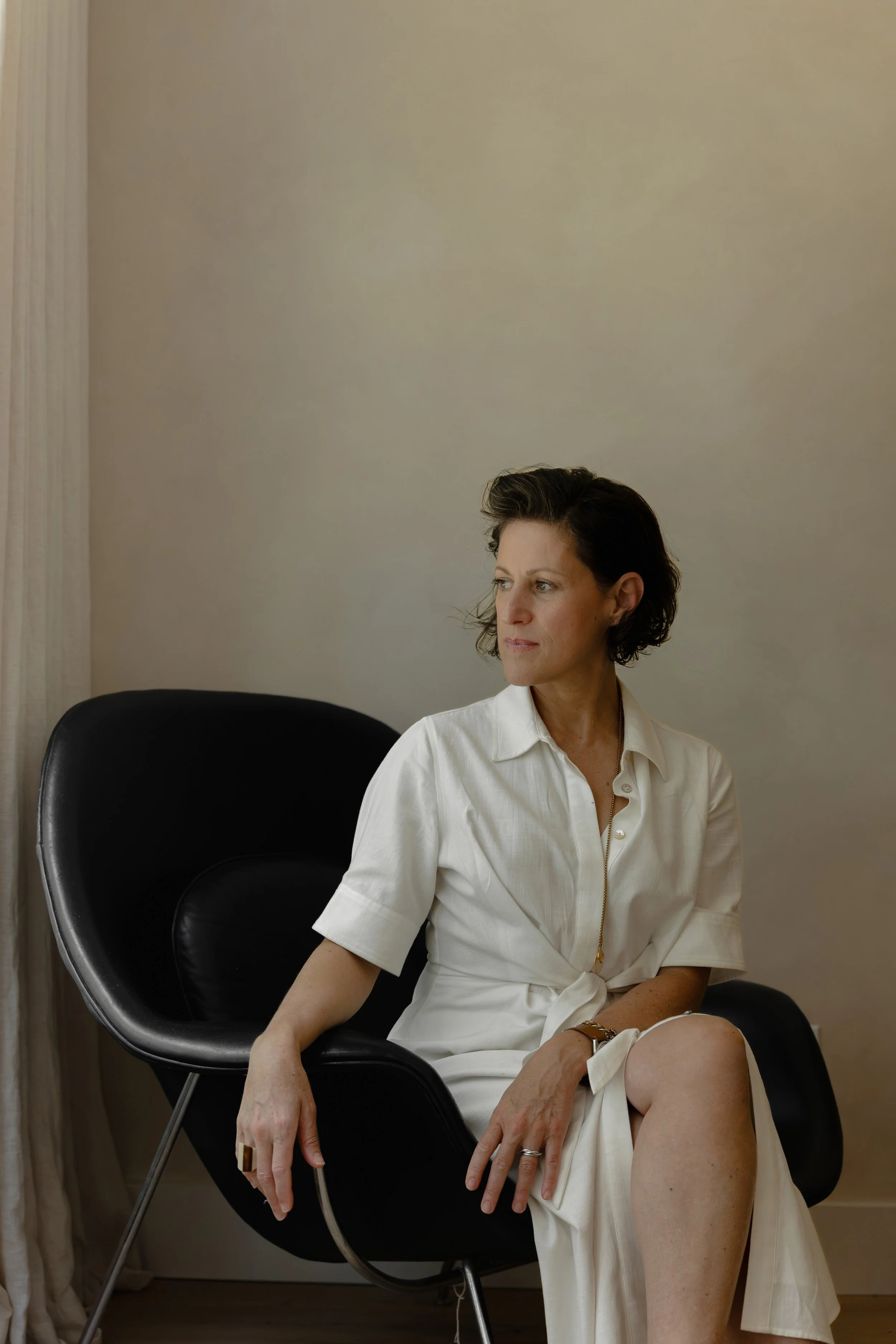 A woman sitting on a black chair next to a beige wall, wearing a cream-colored dress with a zipper, looking to her right with a serious expression.