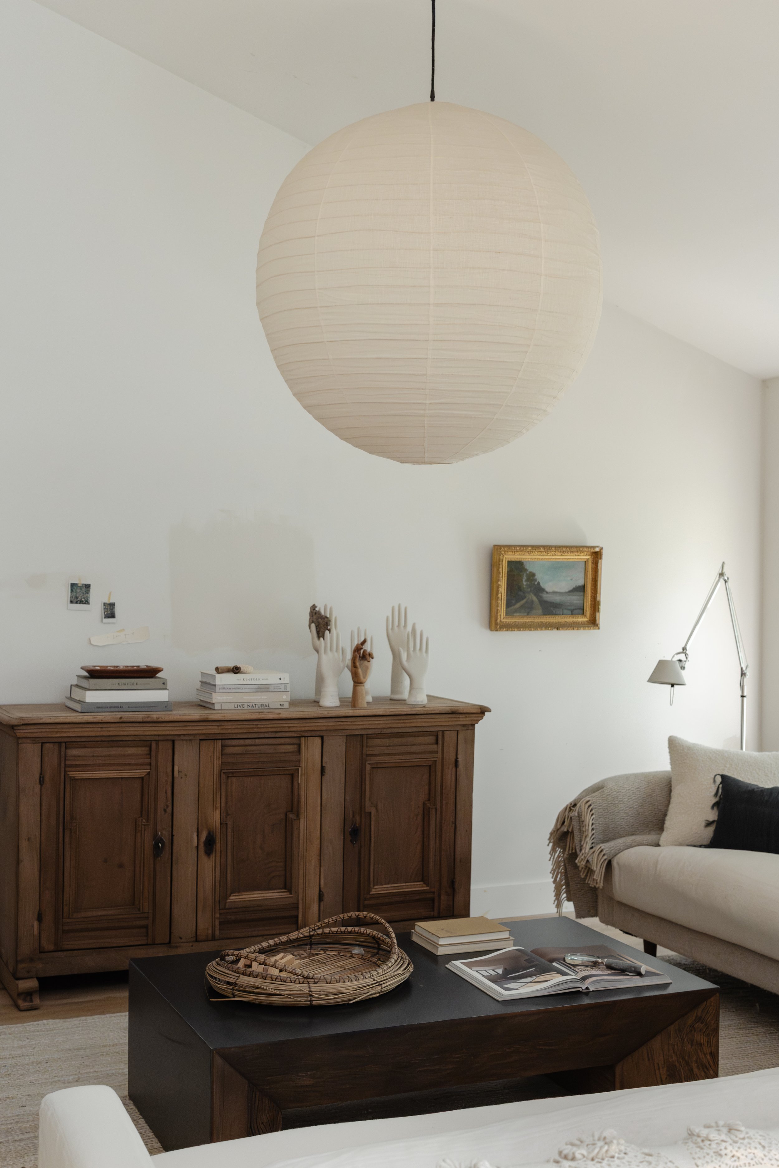 A photo of a living space with high sloped ceilings and a wood vintage cabinet on the wall. The couch sits just right of the cabinet covered in linen fabric. There is linen large spherical lamp hanging in the center and a small painting hanging.