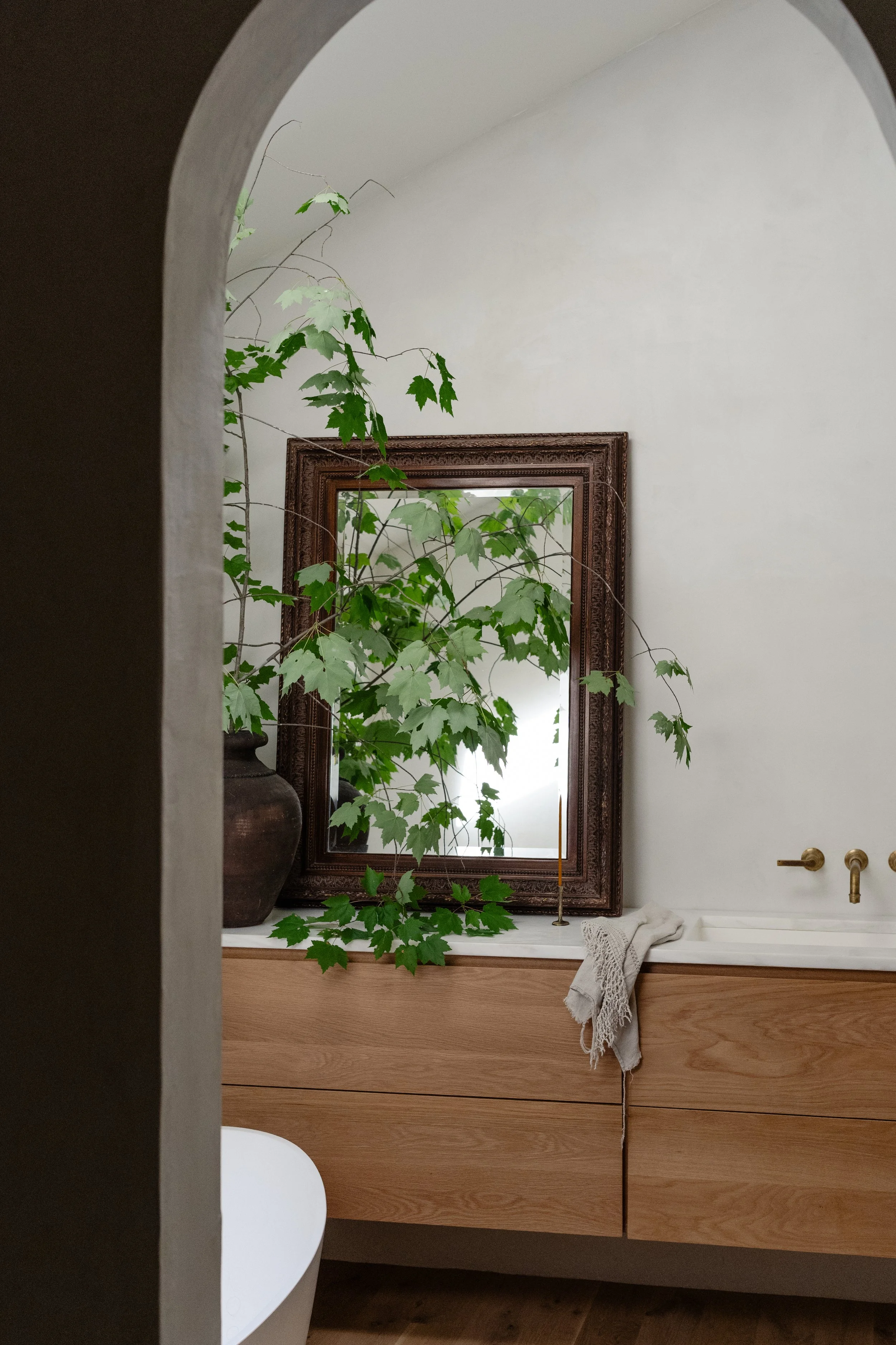 An image of the master bathroom cabinets with a mirror on the countertop and a vase with green limbs in it. There is a small hand towel that rests on the counter.