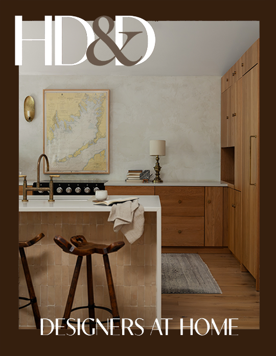Interior view of a modern kitchen with wooden cabinets, a white island with a beige towel, two bar stools, a stovetop, a map on the wall, and a table lamp, with a gray rug on the wooden floor.