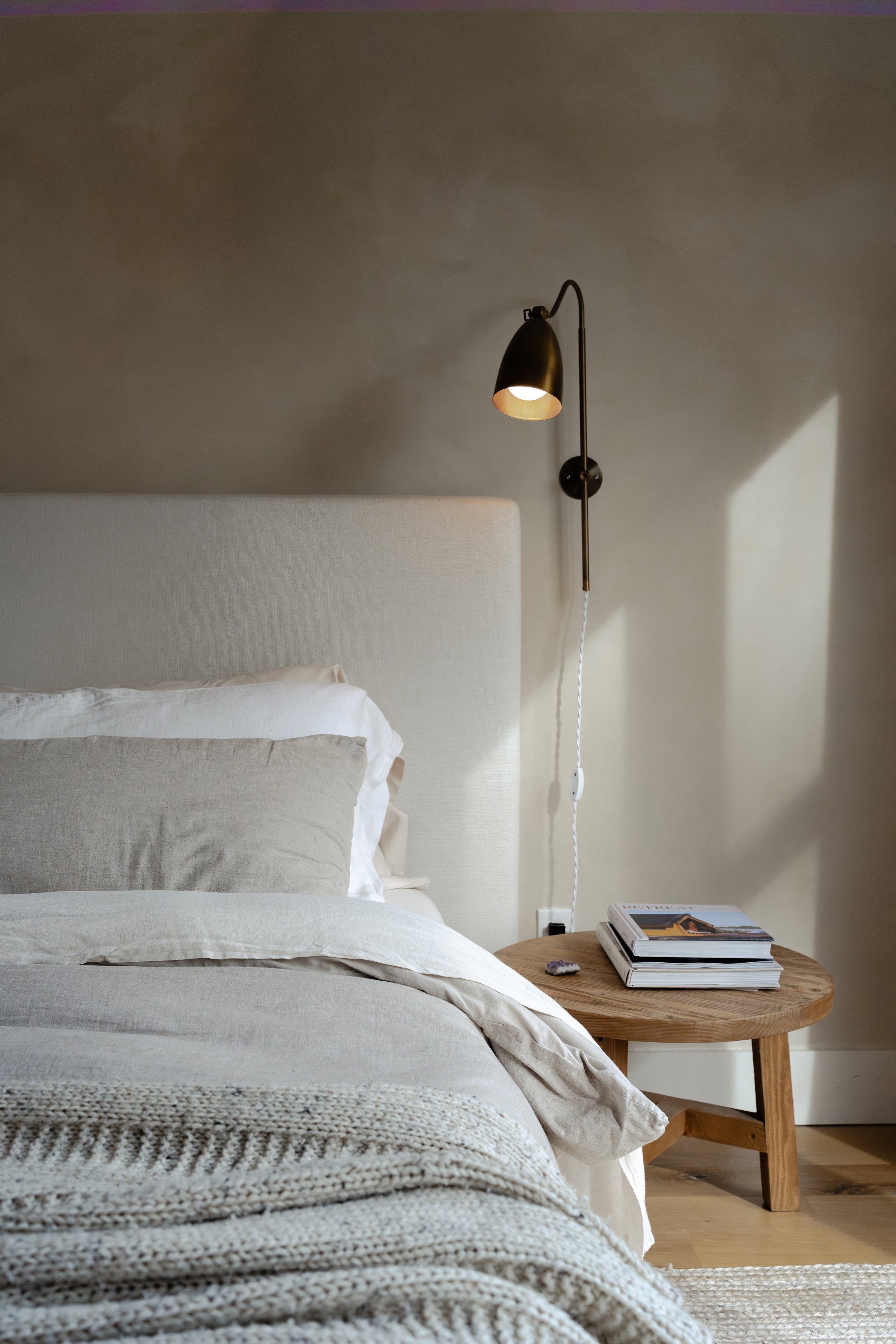 A frontal view of the master bedroom with a linen bedspread and quilt on the bed. The walls are textured painted limewash and the wall metal lamp is turned on.