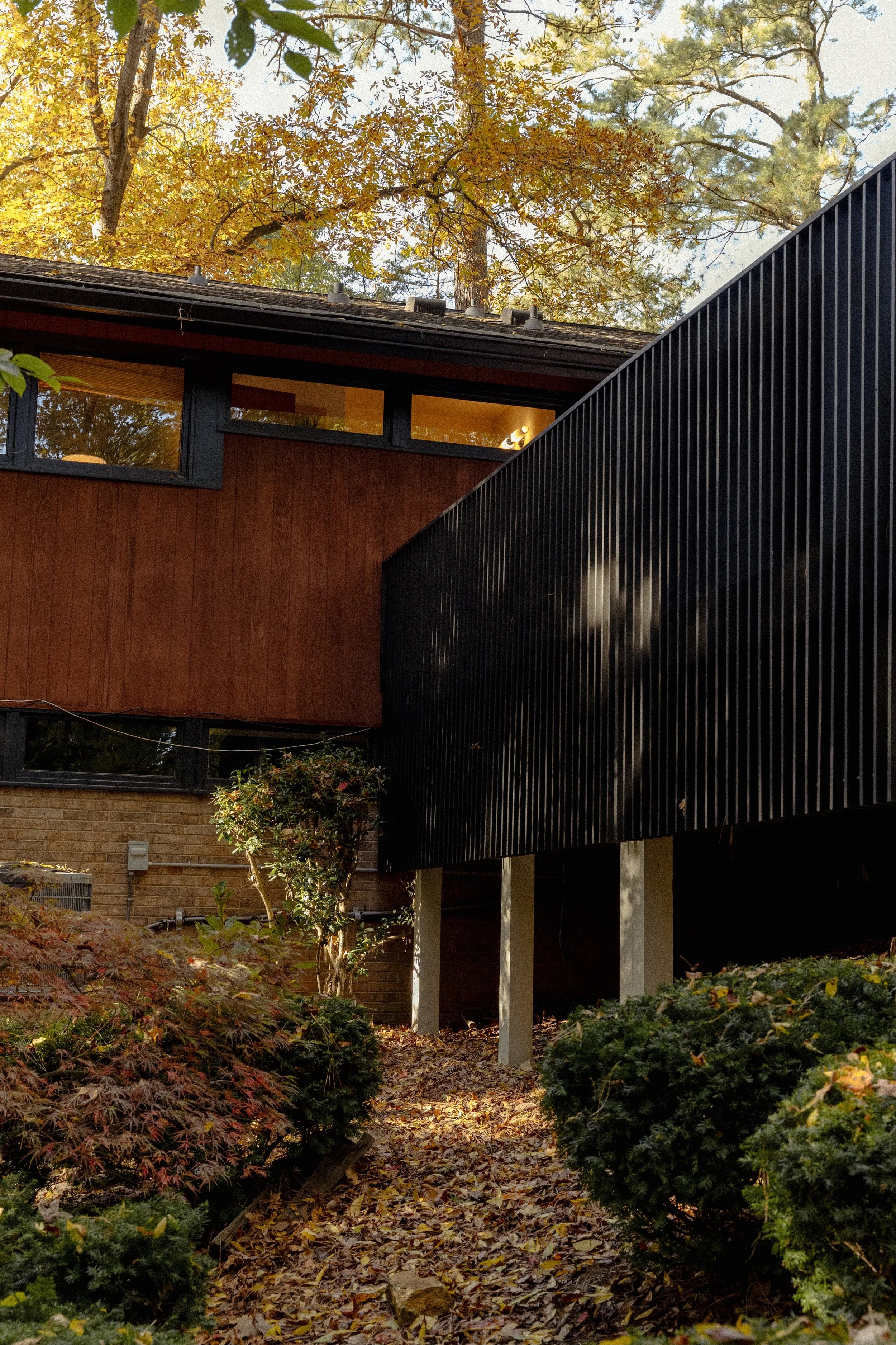 A shot of a midcentury modern house with a deck with vertical slats. The deck is situated inside the garden with some bushes and a small maple in the foreground.
