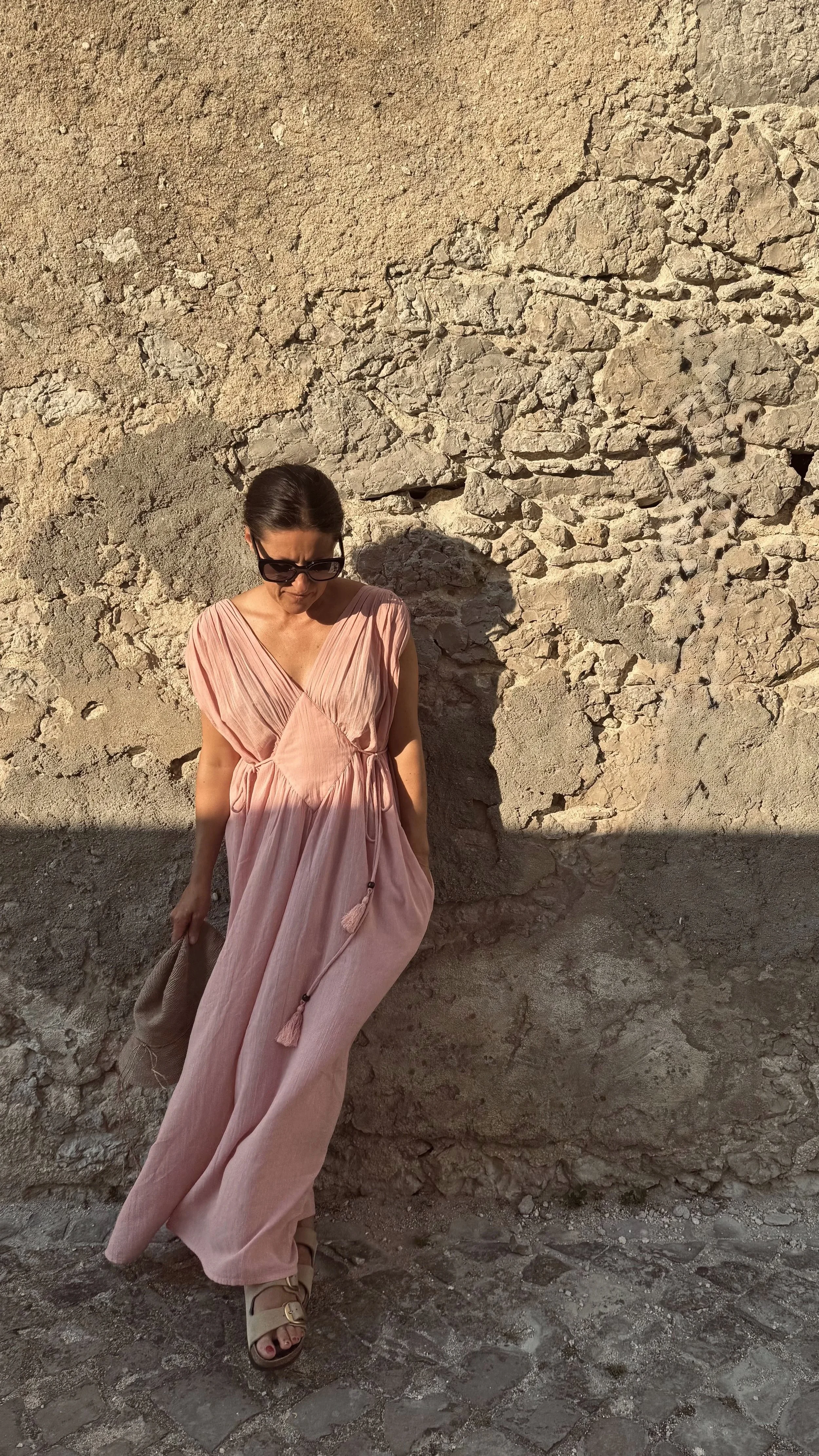 A woman with dark hair, wearing sunglasses and a flowing pink dress with tassels, standing next to a textured stone wall.