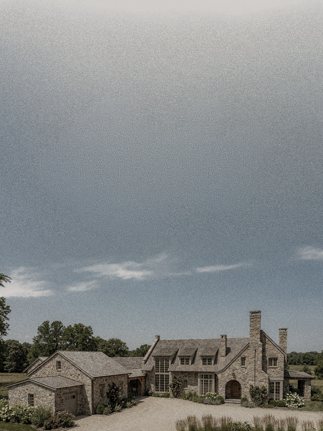 A large stone house with multiple gabled roofs, chimneys, and tall windows, set in a landscaped yard with shrubs and trees under a cloudy sky.