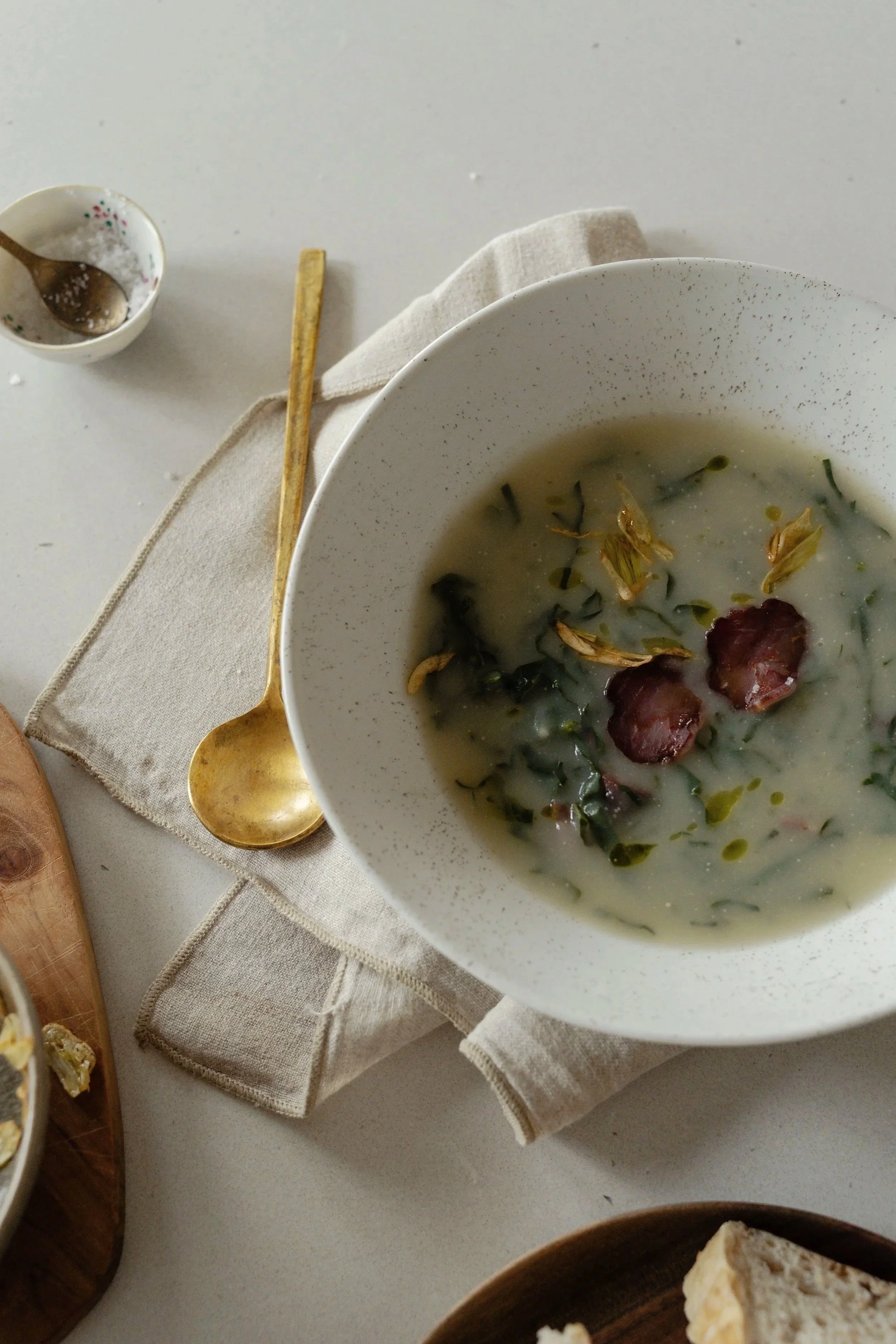 A bowl of soup with chopped herbs and toppings, a small bowl of salt with a spoon, and a gold-colored spoon on a beige napkin.