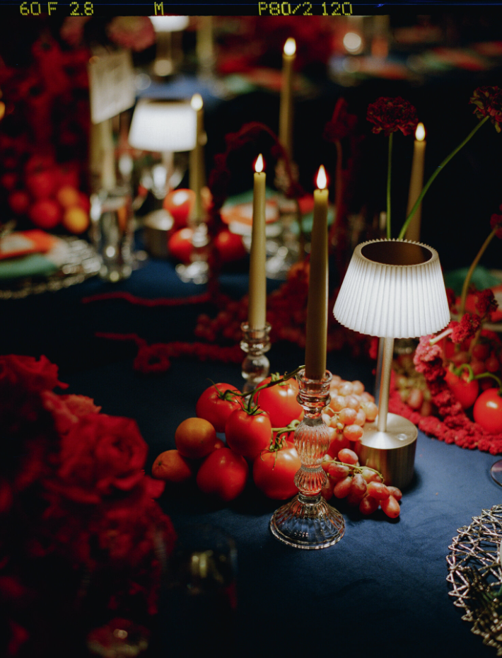 A table decorated with red tomatoes, grapes, floral arrangements, candle holders with lit and unlit candles, and a small white lamp with a pleated shade, in a dimly lit setting.