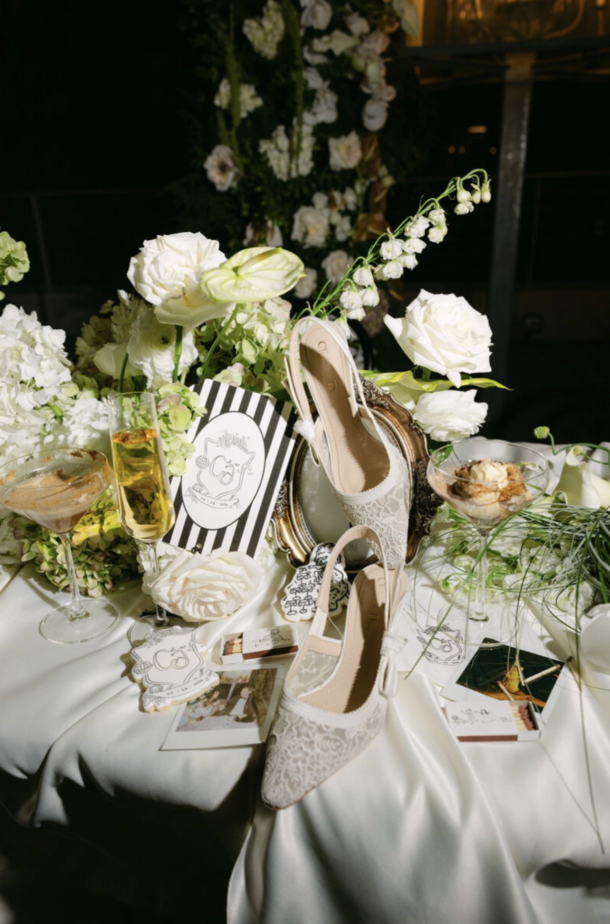 A decorated table with white floral arrangements, lace high-heeled shoes, glasses of champagne and dessert, and a framed mirror at a wedding reception.
