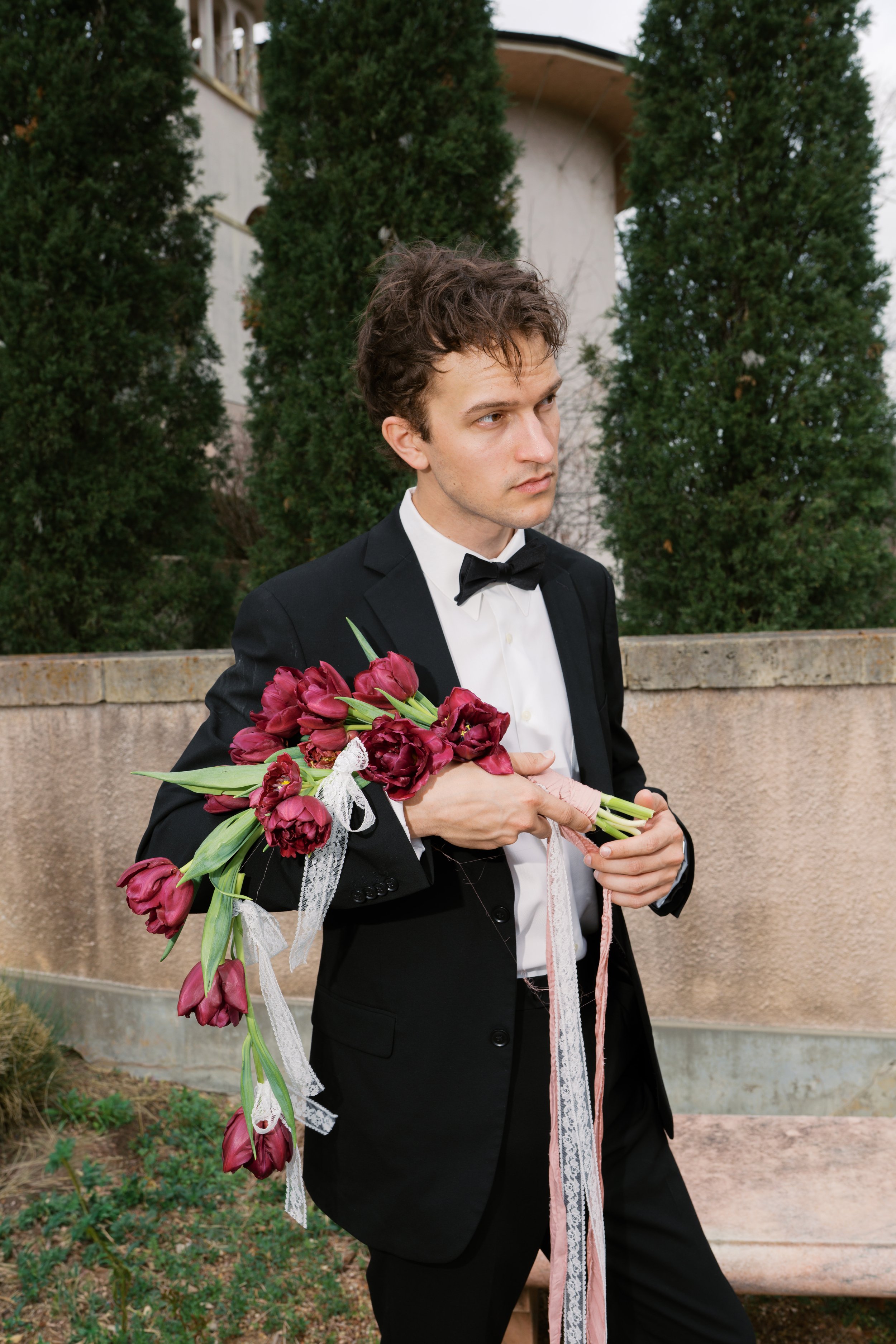 Man in black tuxedo holding a bouquet of red tulips, standing outdoors with trees and a concrete wall in the background.