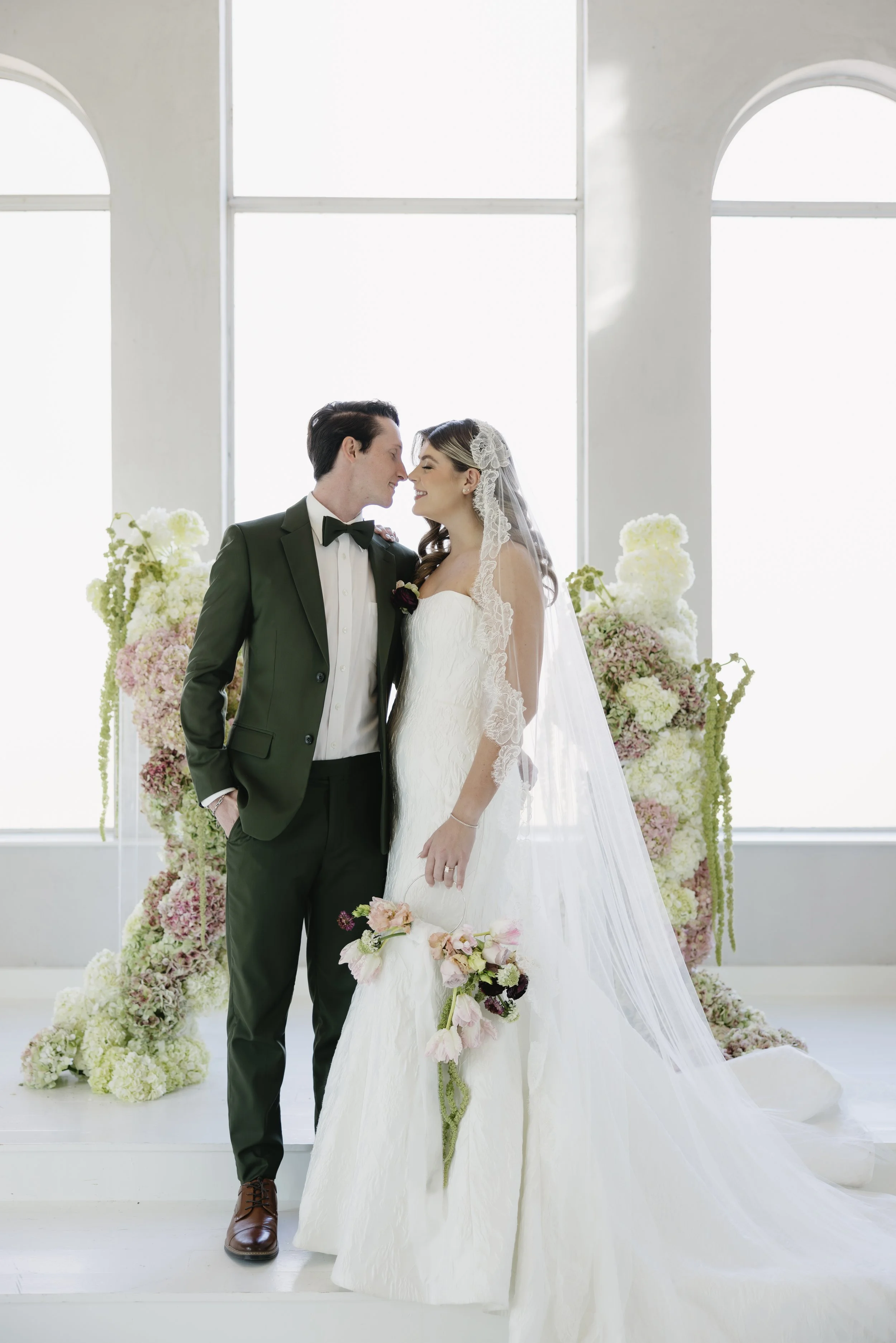 A bride in a white wedding dress holding a bouquet of flowers, and a groom in a dark suit with a bowtie, standing close together with foreheads touching in a bright, flower-decorated venue.