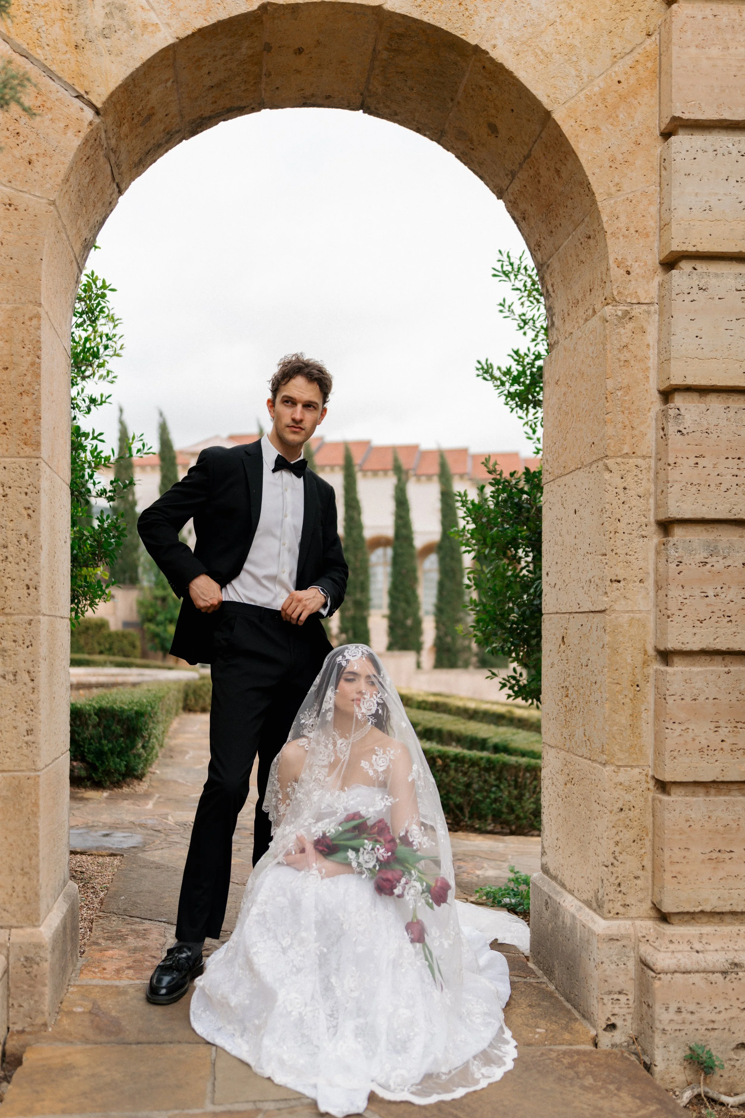 A bride in a white wedding dress and veil sitting outdoors holding a bouquet of red and white flowers, with a groom in a black tuxedo standing beside her under a stone archway, with a garden and cypress trees in the background.