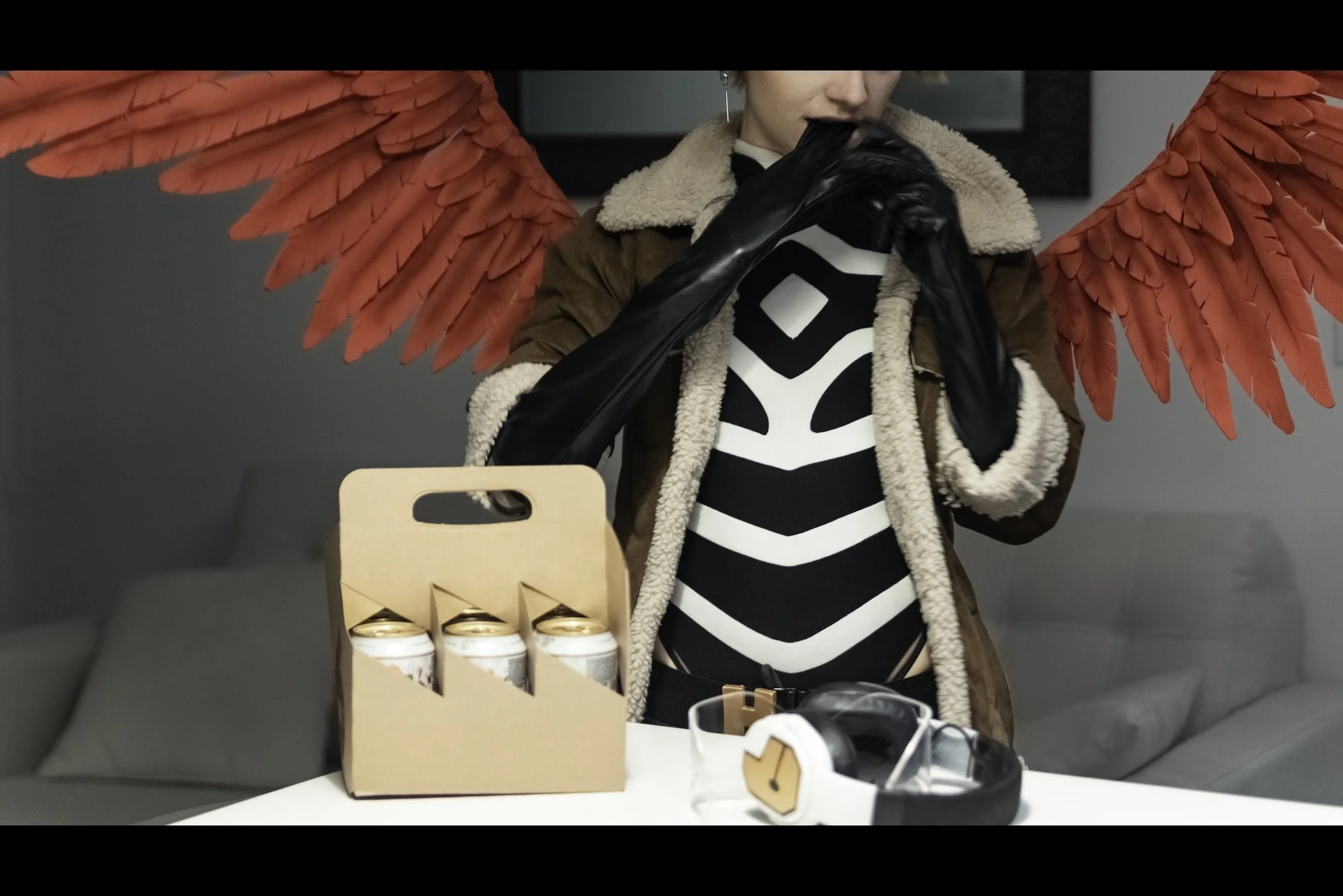 View of the six pack set on the table, with Hawks' headset and goggles; in the background, Hawks pulls off one of his gloves with his teeth.