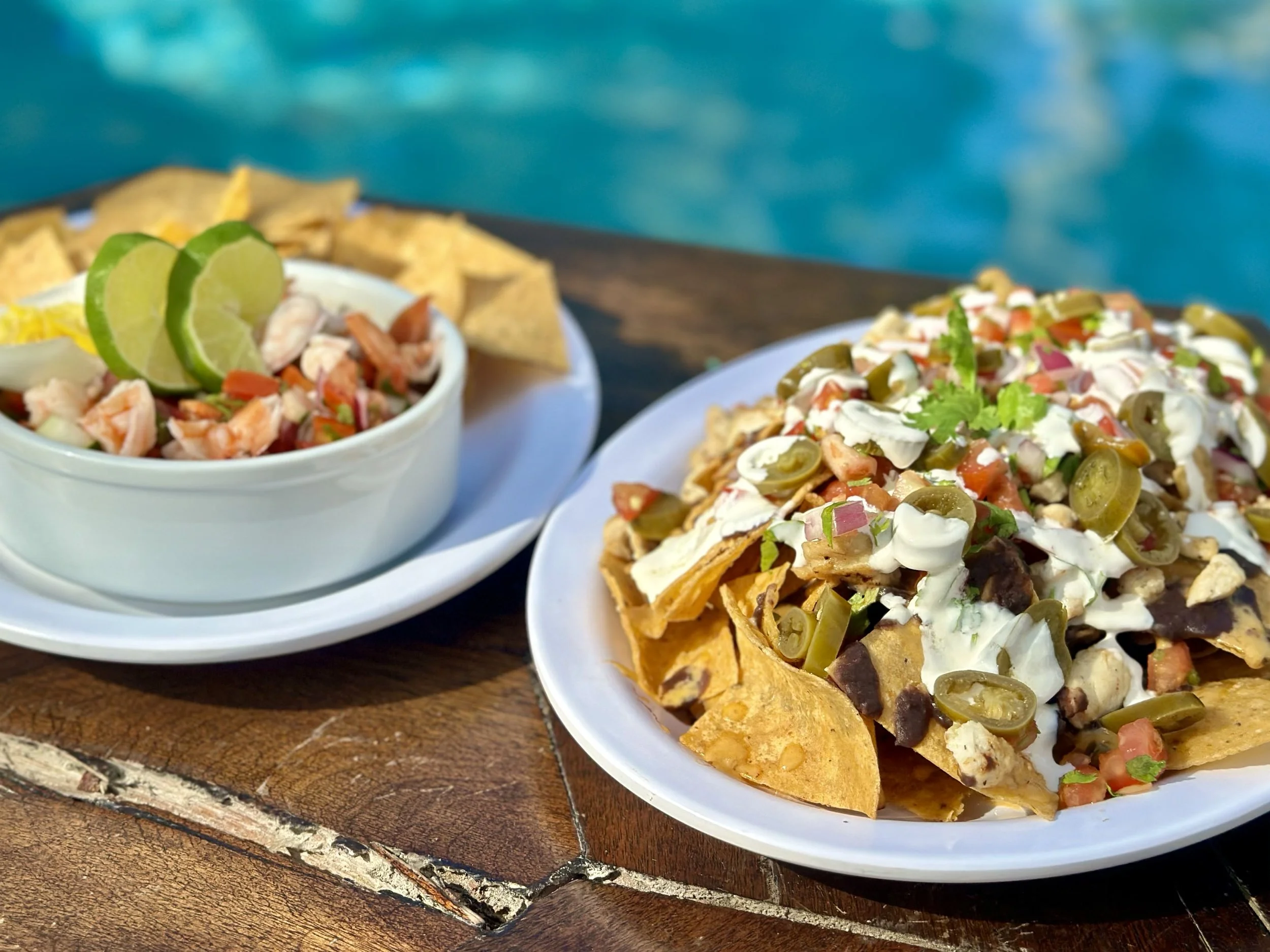Plate of nachos topped with chicken, sour cream, jalapeños, tomatoes, and cilantro, next to a bowl of shrimp cocktail garnished with lime wedges and tortilla chips, on a wooden table with a blue background.