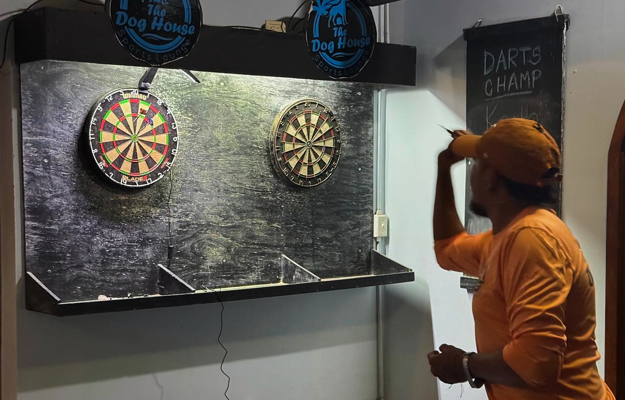 A man in an orange shirt and cap playing darts at a darts game in a bar or pub. Two dartboards are mounted on the wall, and a chalkboard sign reads 'Darts Champ'.