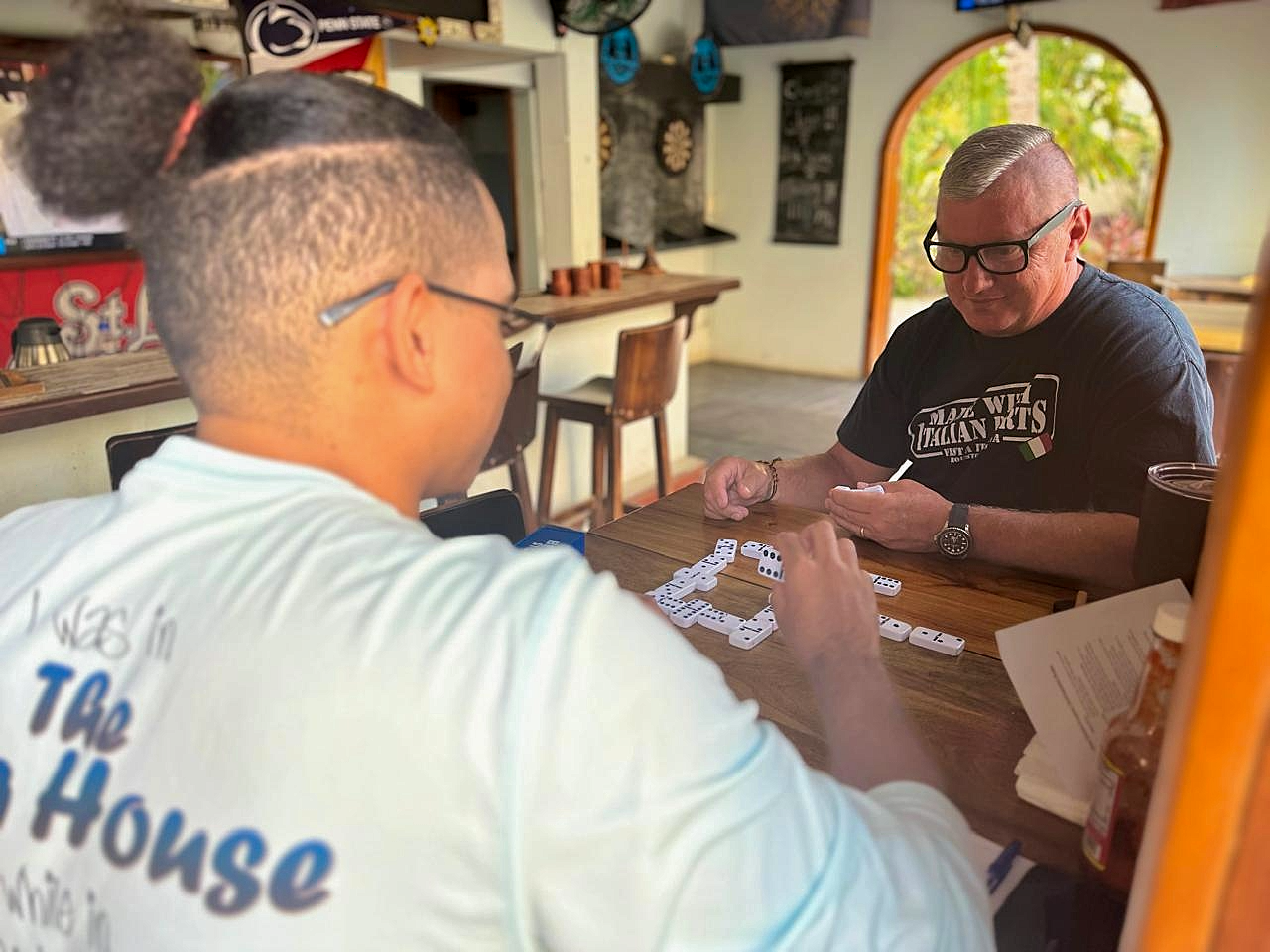 Two men are playing dominoes at a wooden table in a cozy, brightly-lit cafe or restaurant. One man has short blond hair and black glasses, while the other has dark hair tied in a bun and is wearing a white shirt with blue text on the back. The backgr