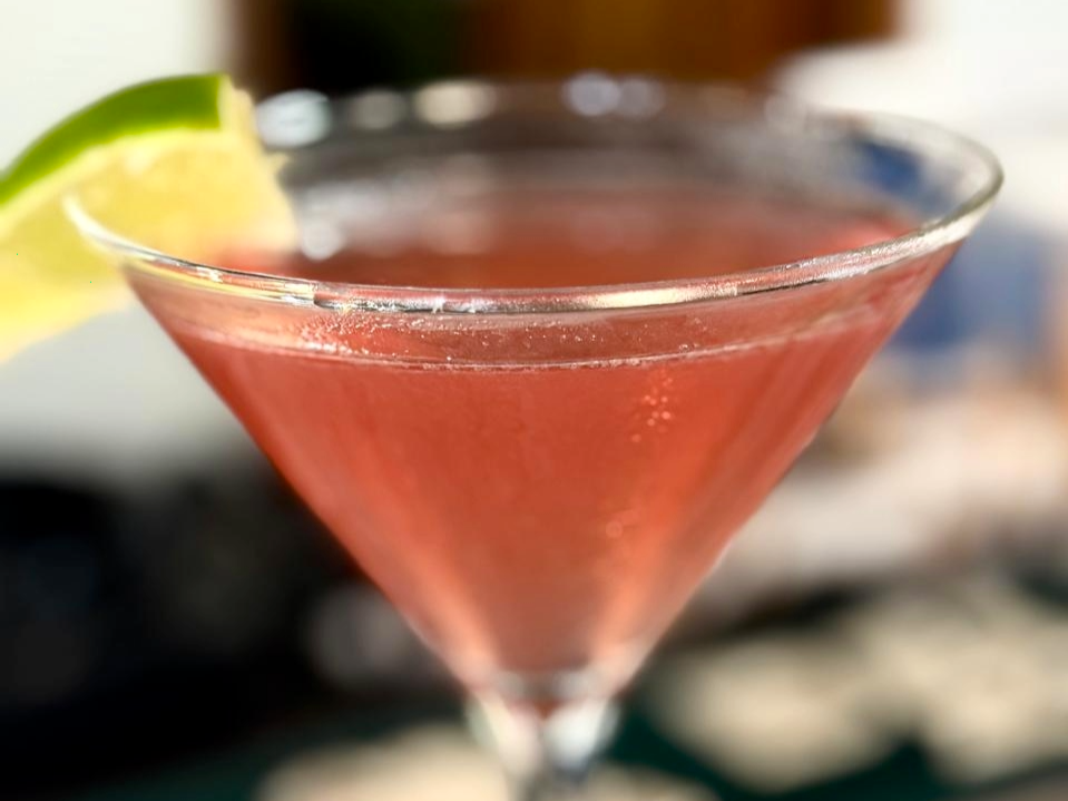 A pink cocktail served in a martini glass with a lemon and lime wedge garnish.