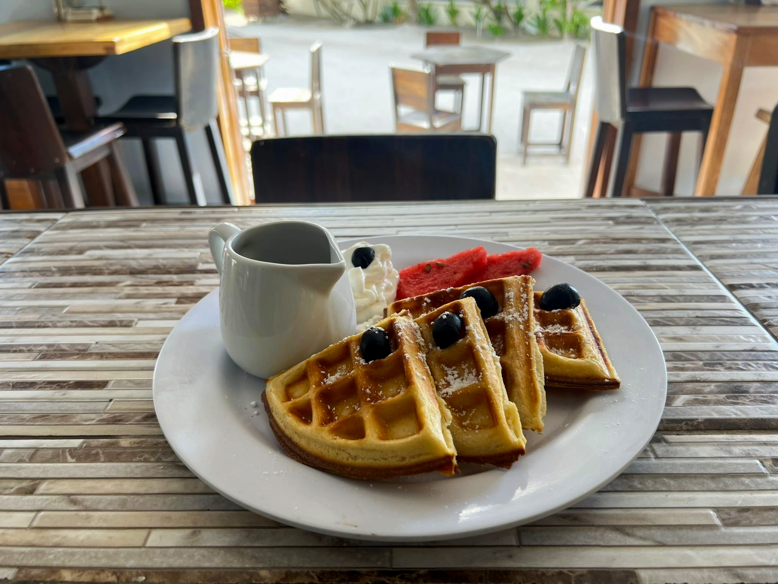 Plate with Belgian waffles topped with blueberries, served with watermelon slices, whipped cream, and a small pitcher of syrup on a wooden table at a café.