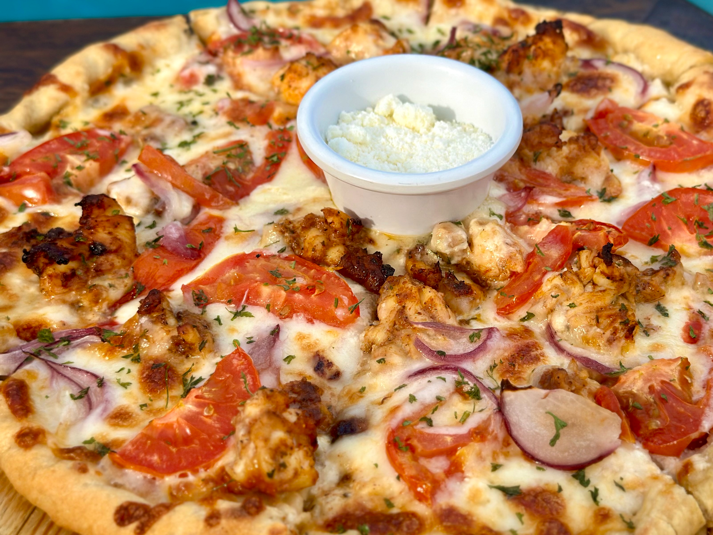 Close-up of a pizza with cheese, tomatoes, onions, grilled chicken, and herbs, with a small bowl of grated cheese or Parmesan in the center.